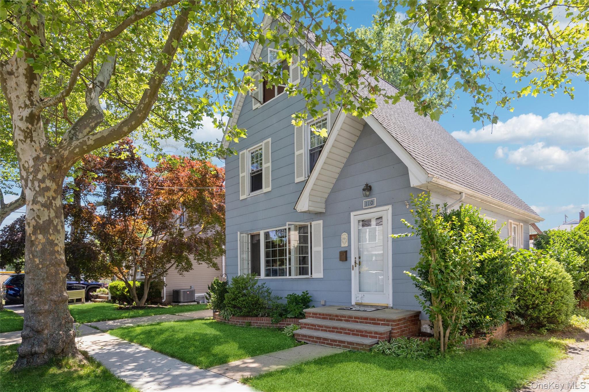 Blue-gray two-story house with white trim, brick front steps, and a large shade tree over the lawn.