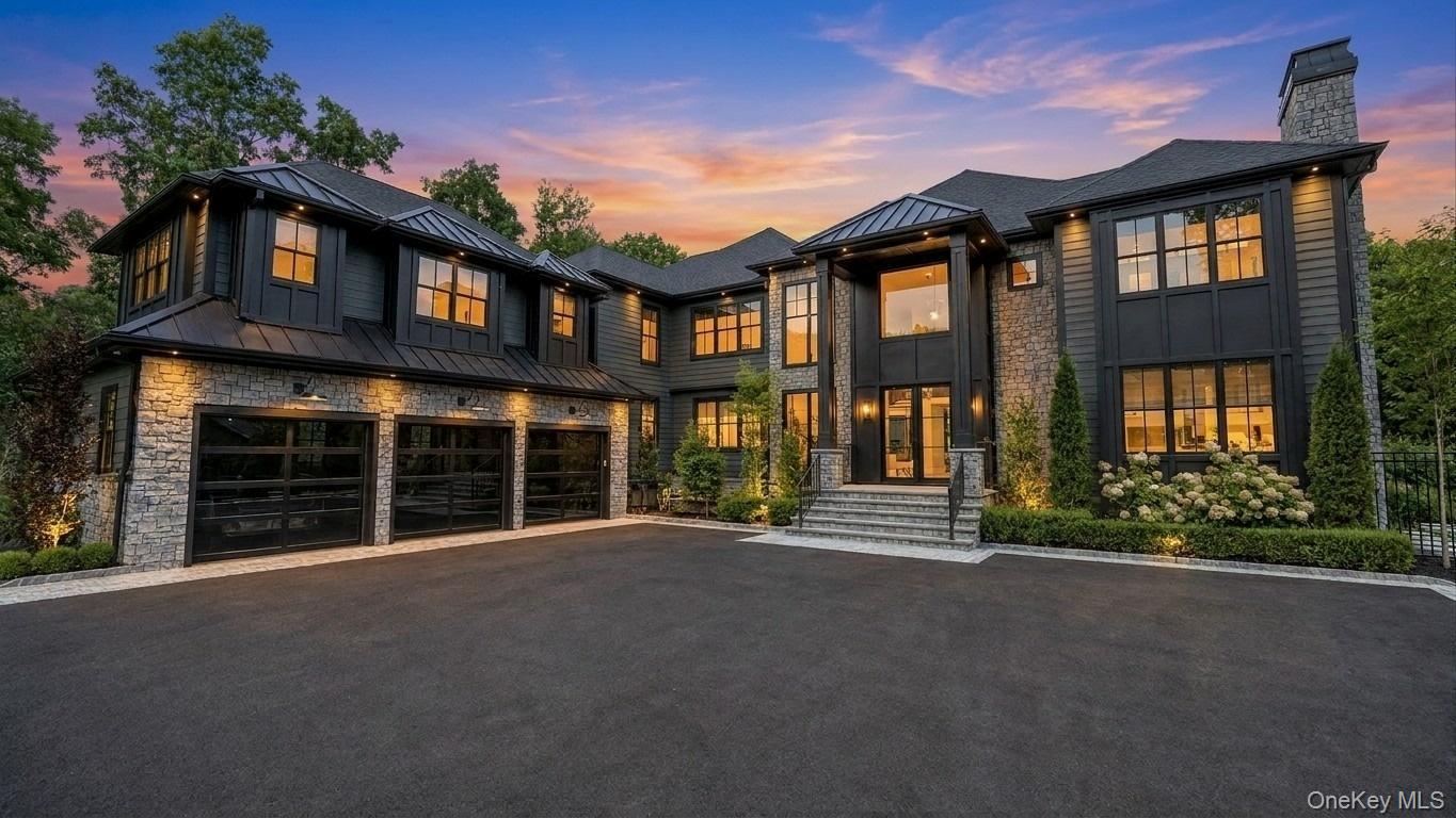 Two-story stone-and-dark-siding home with a three-car garage, glowing windows, and a sunset sky.