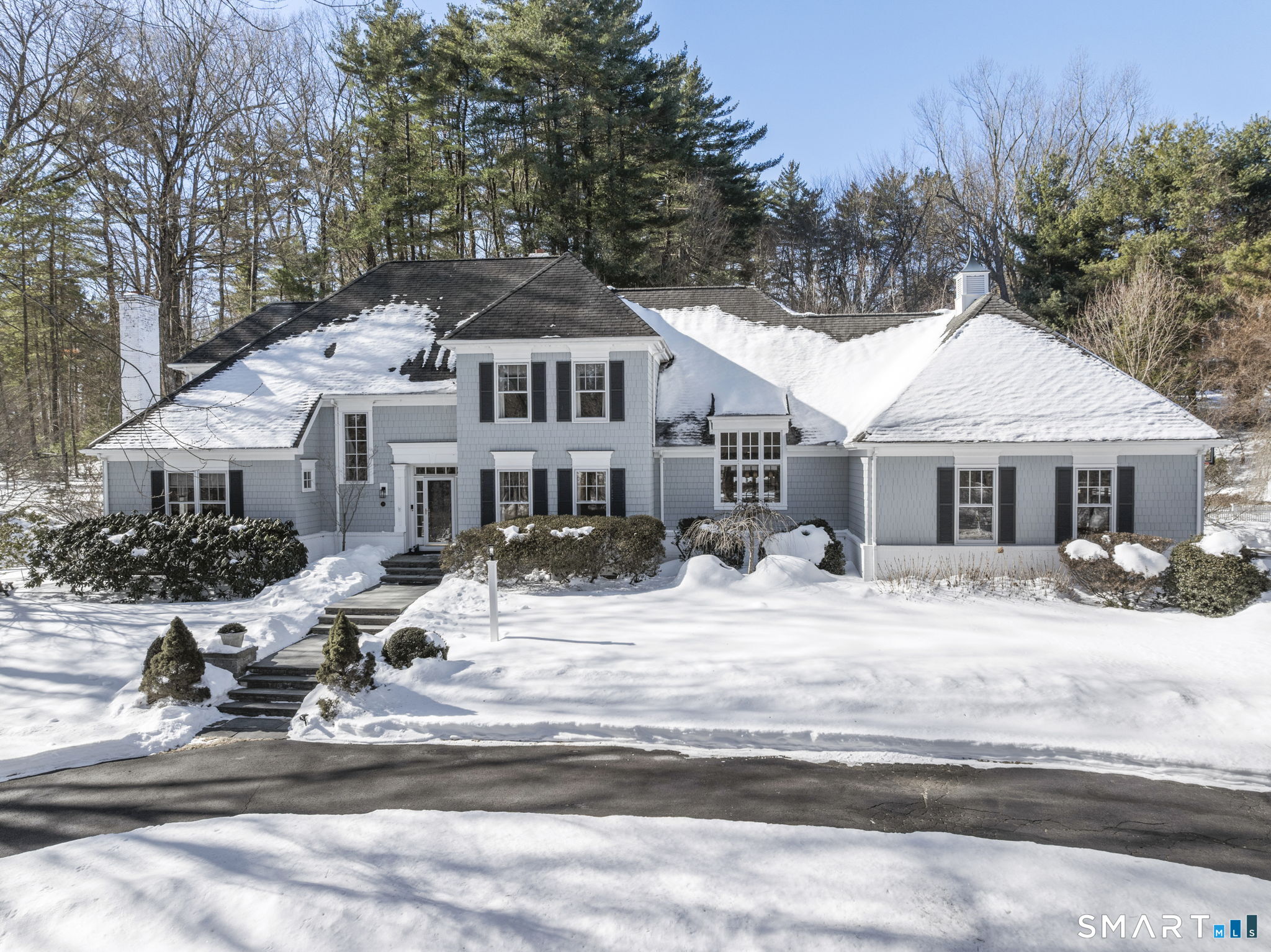 Gray shingle-sided home with dark shutters, photographed in winter with snow covering the yard and roof.