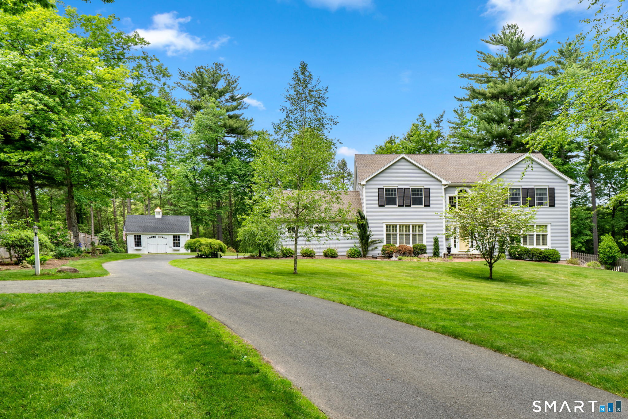 White two-story house with a curved driveway, detached garage, and wooded surroundings.