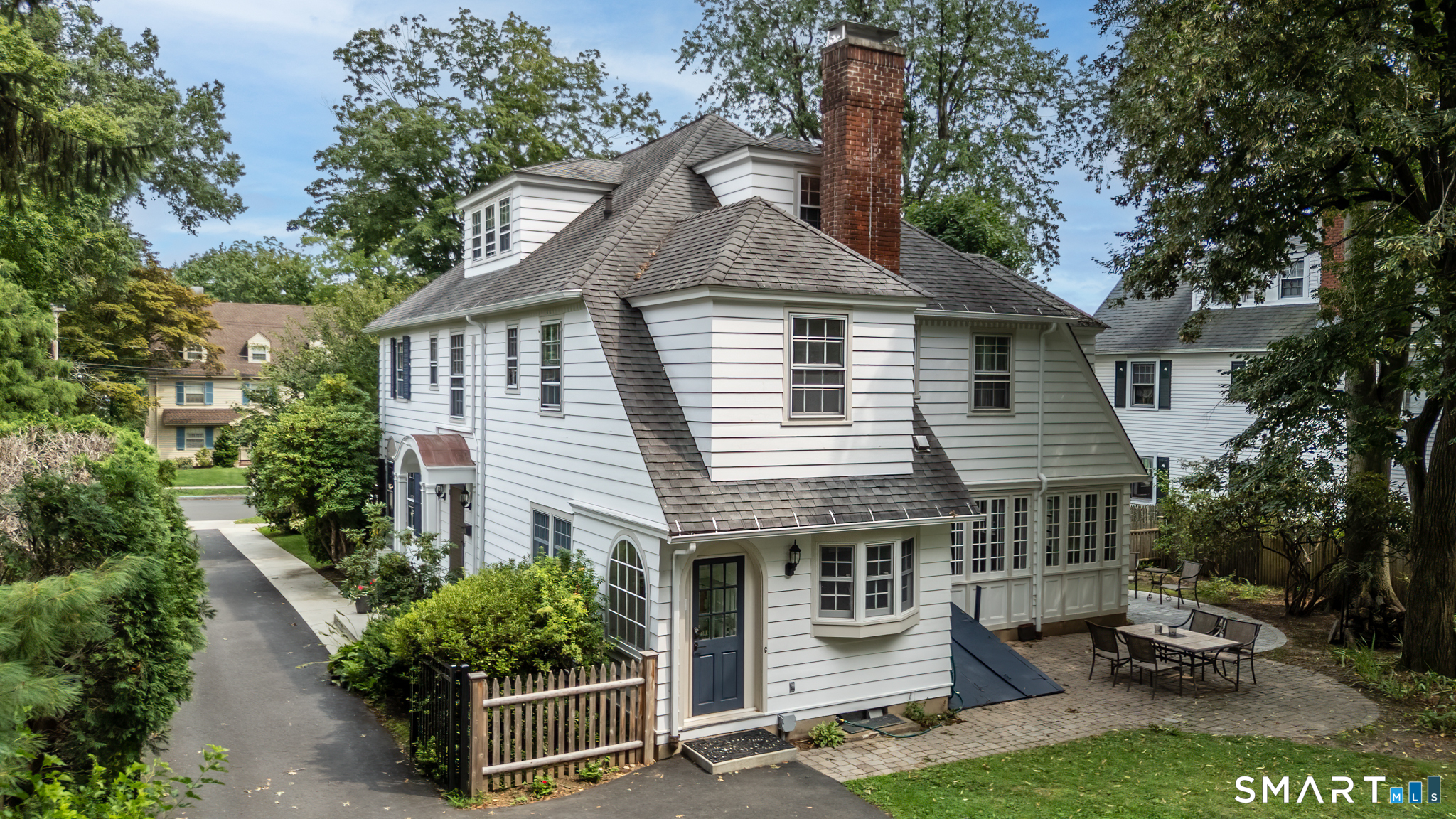 White multi-story house with a tall brick chimney, blue front door, and shaded stone patio.