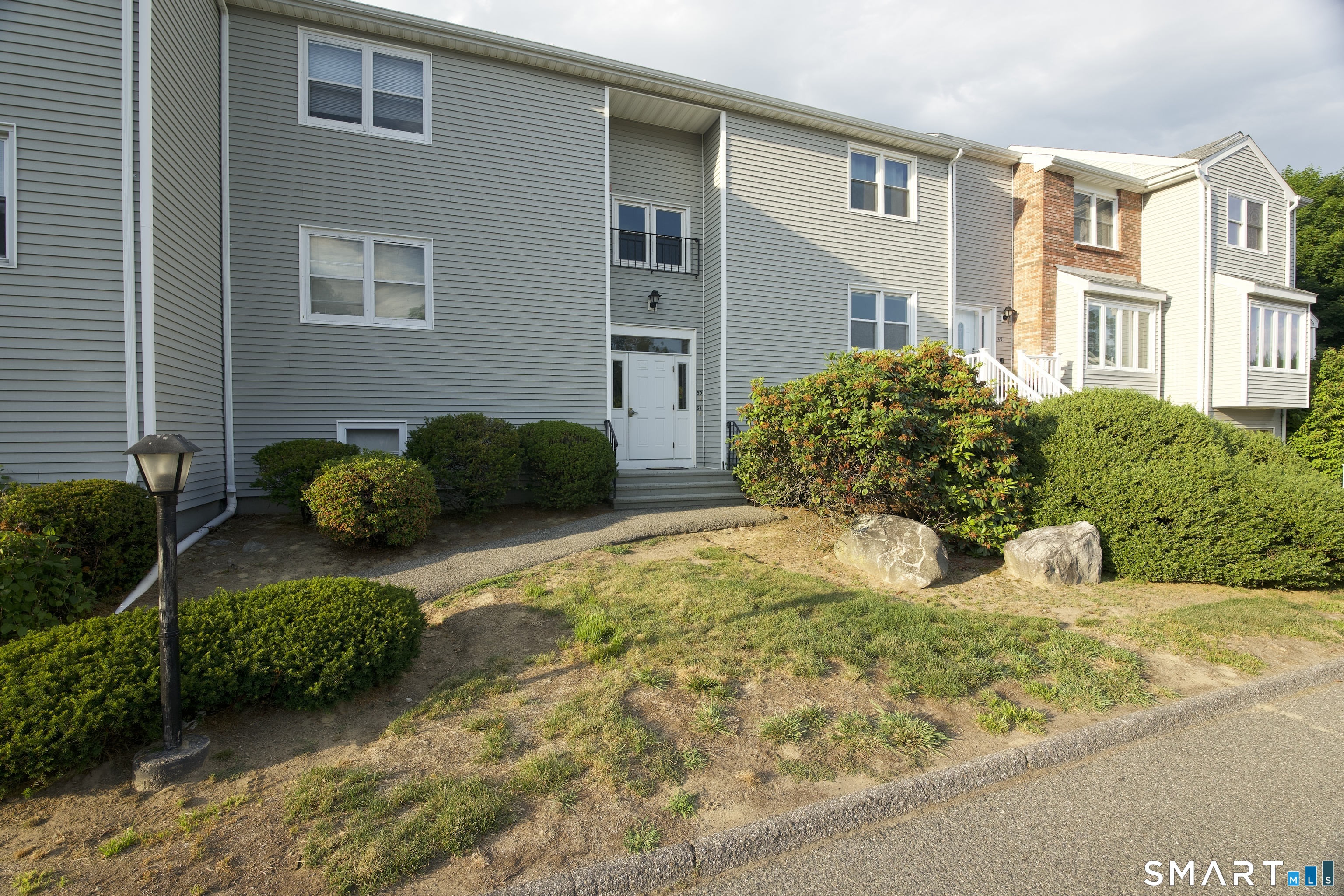 Gray-sided townhouse with a white front door, balcony, and landscaped shrubs.