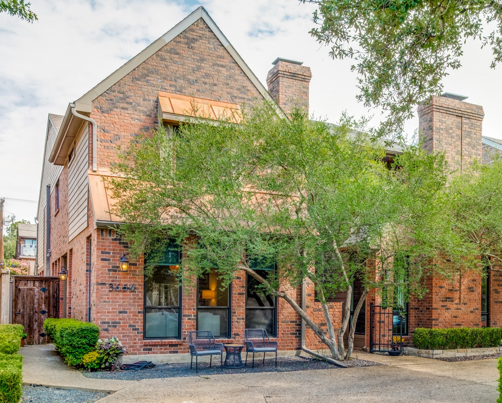 Brick home with copper awnings, mature trees, and a small patio with outdoor chairs.