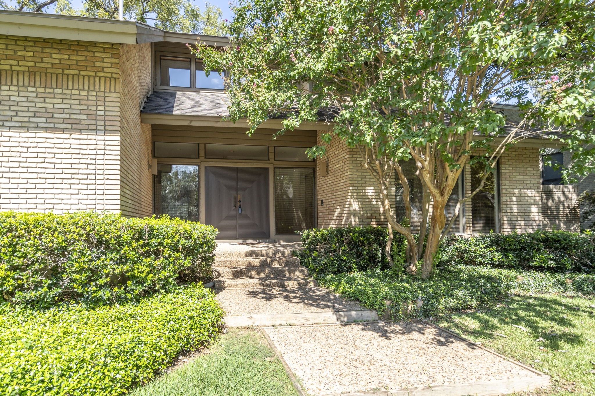 Brick house entrance with a recessed double door, landscaped walkway, and flowering tree.