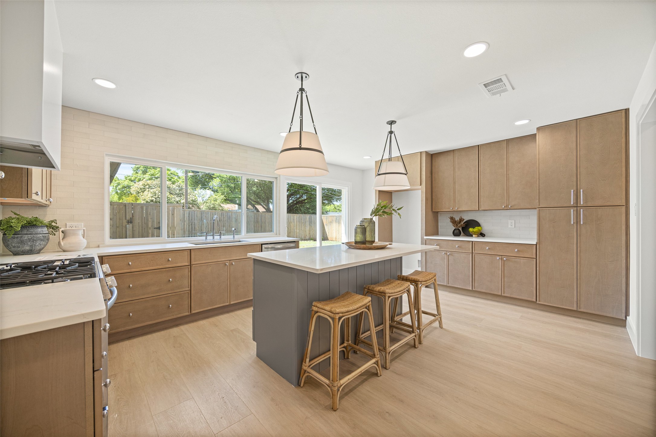 Kitchen with wood cabinets, gray island and woven stools beside large windows overlooking the yard.
