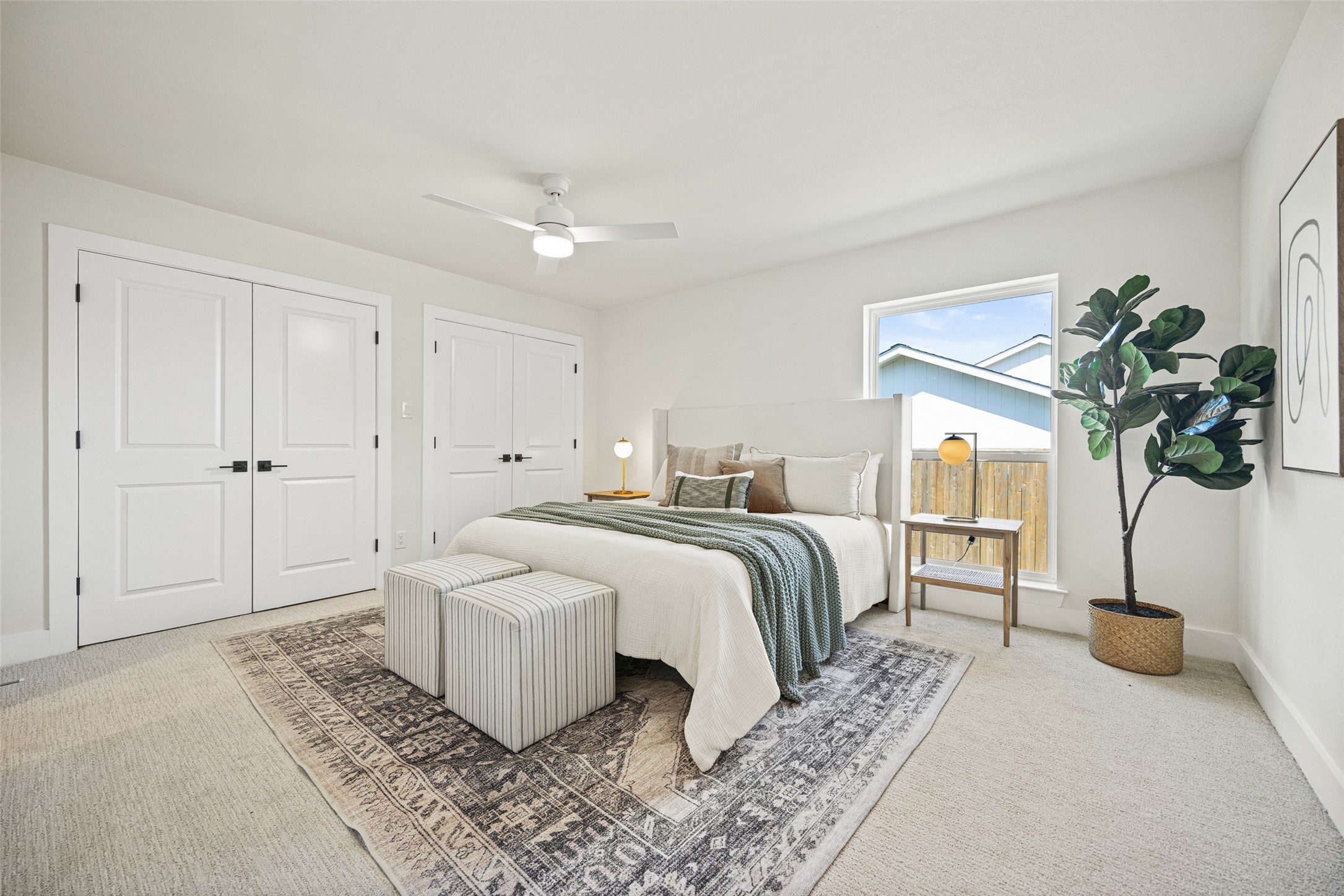 Staged bedroom with a white bed, green throw, striped benches, patterned rug, and large window.