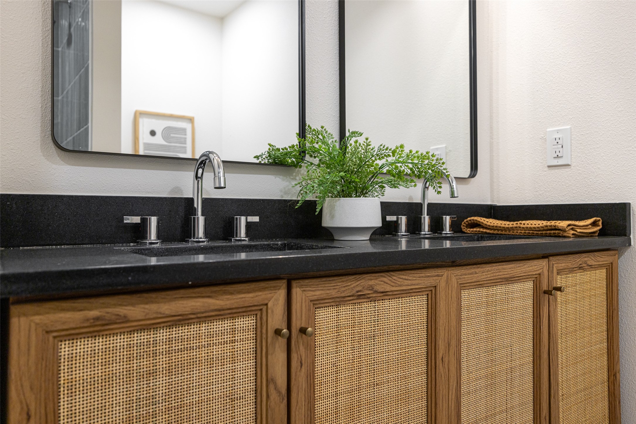 Bathroom vanity with double chrome faucets, dark countertop, and warm wood cabinets with woven fronts.