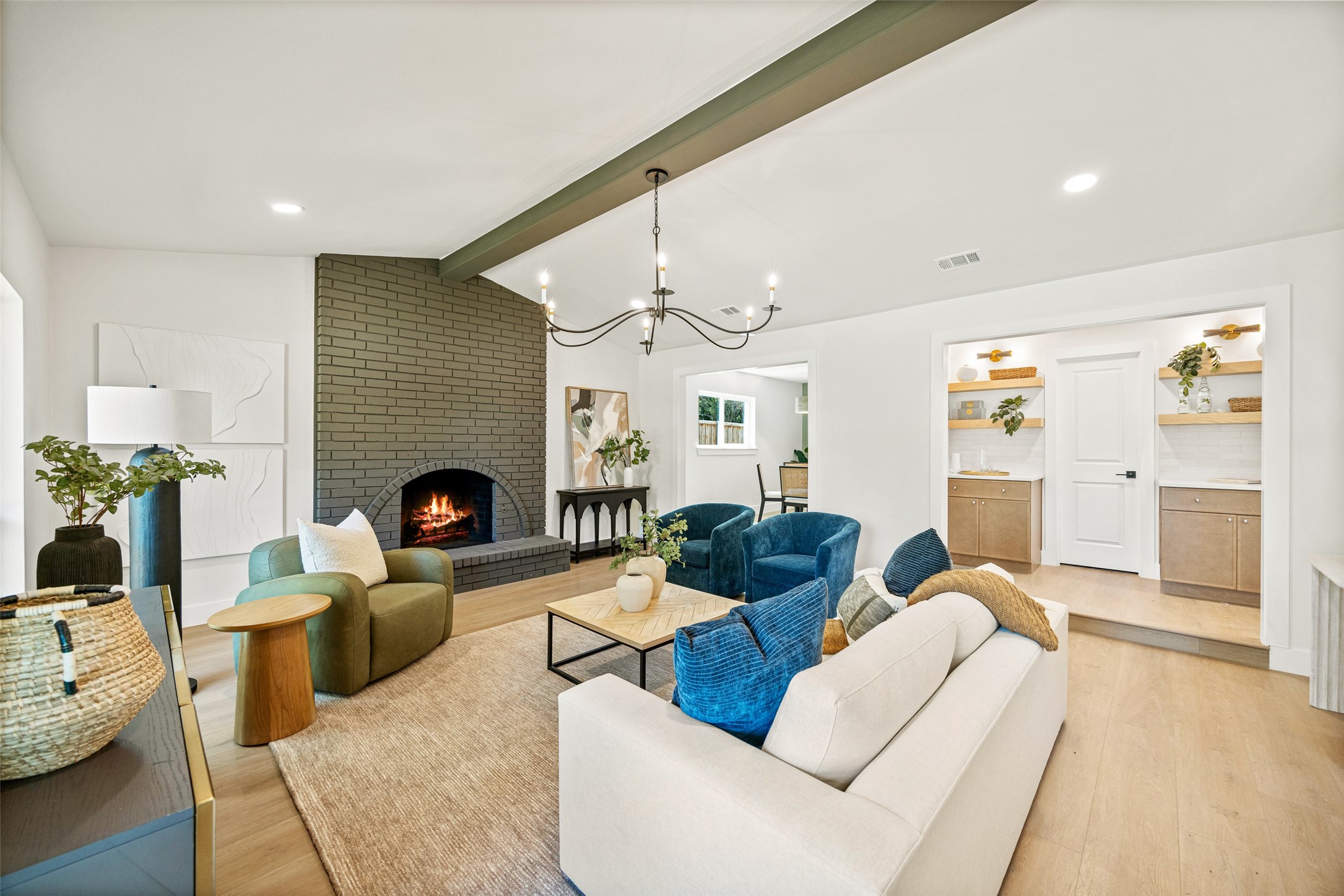 Staged living room with a lit brick fireplace, exposed beam, and blue and green chairs.