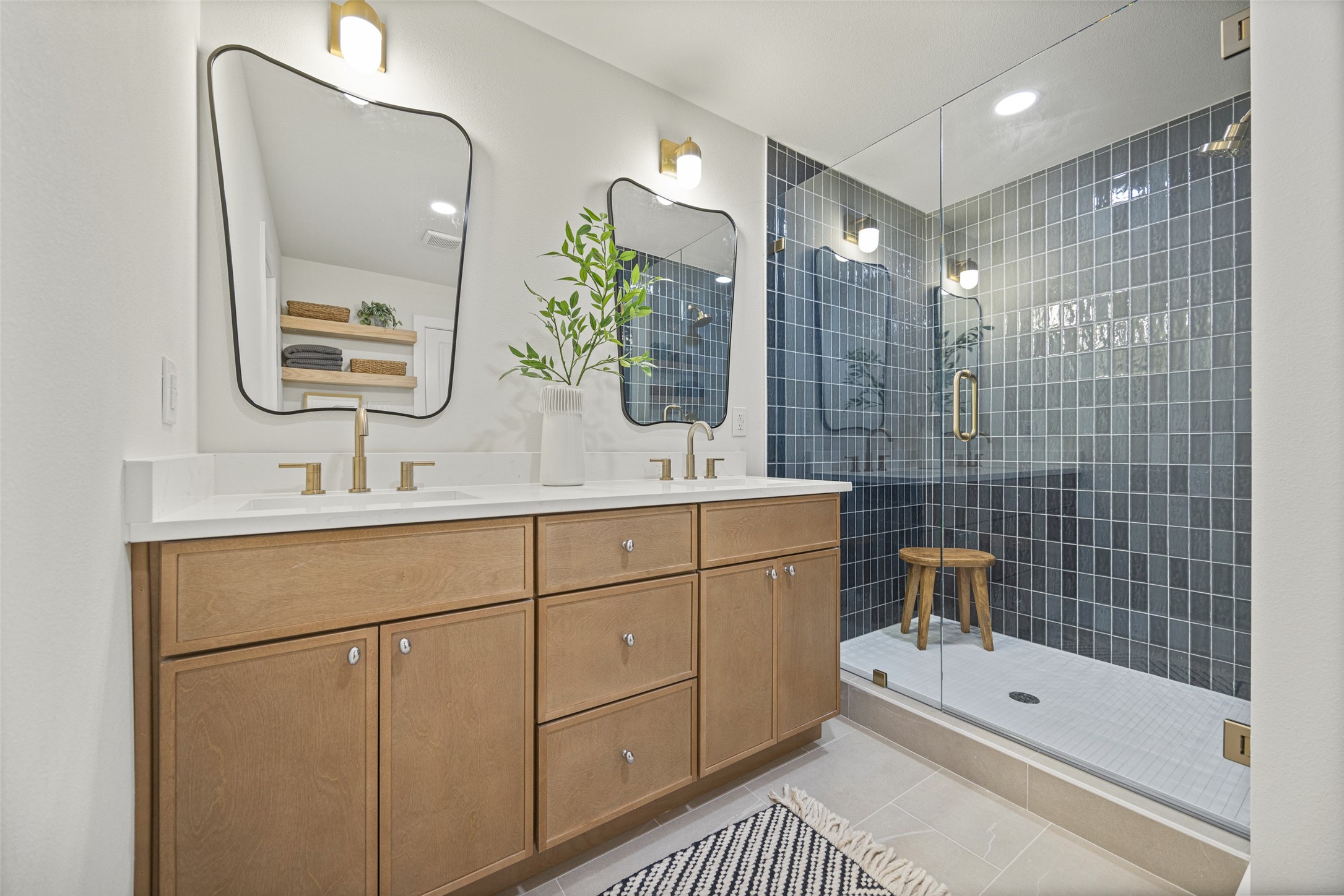 Bathroom with a light wood double vanity and a glass shower lined with dark blue-gray tile.