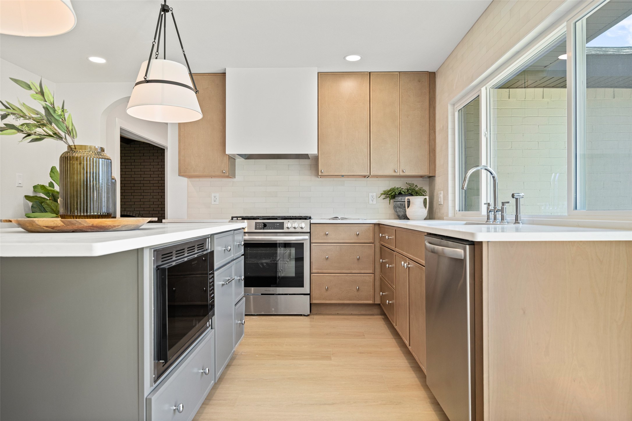 Kitchen with light wood cabinetry, stainless appliances, and a sink beneath large windows.