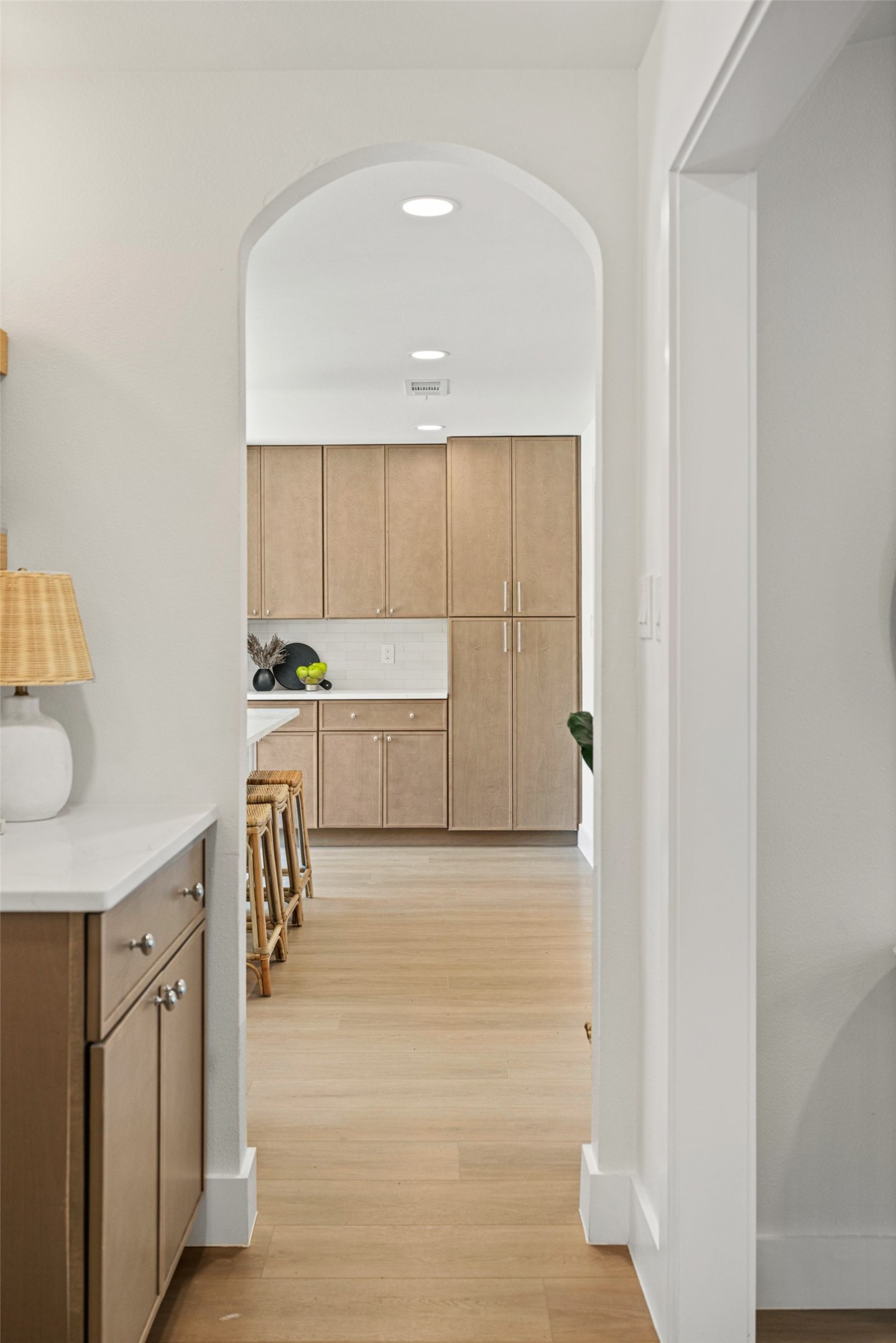 Kitchen with light wood cabinets and woven stools, seen through an arched doorway.
