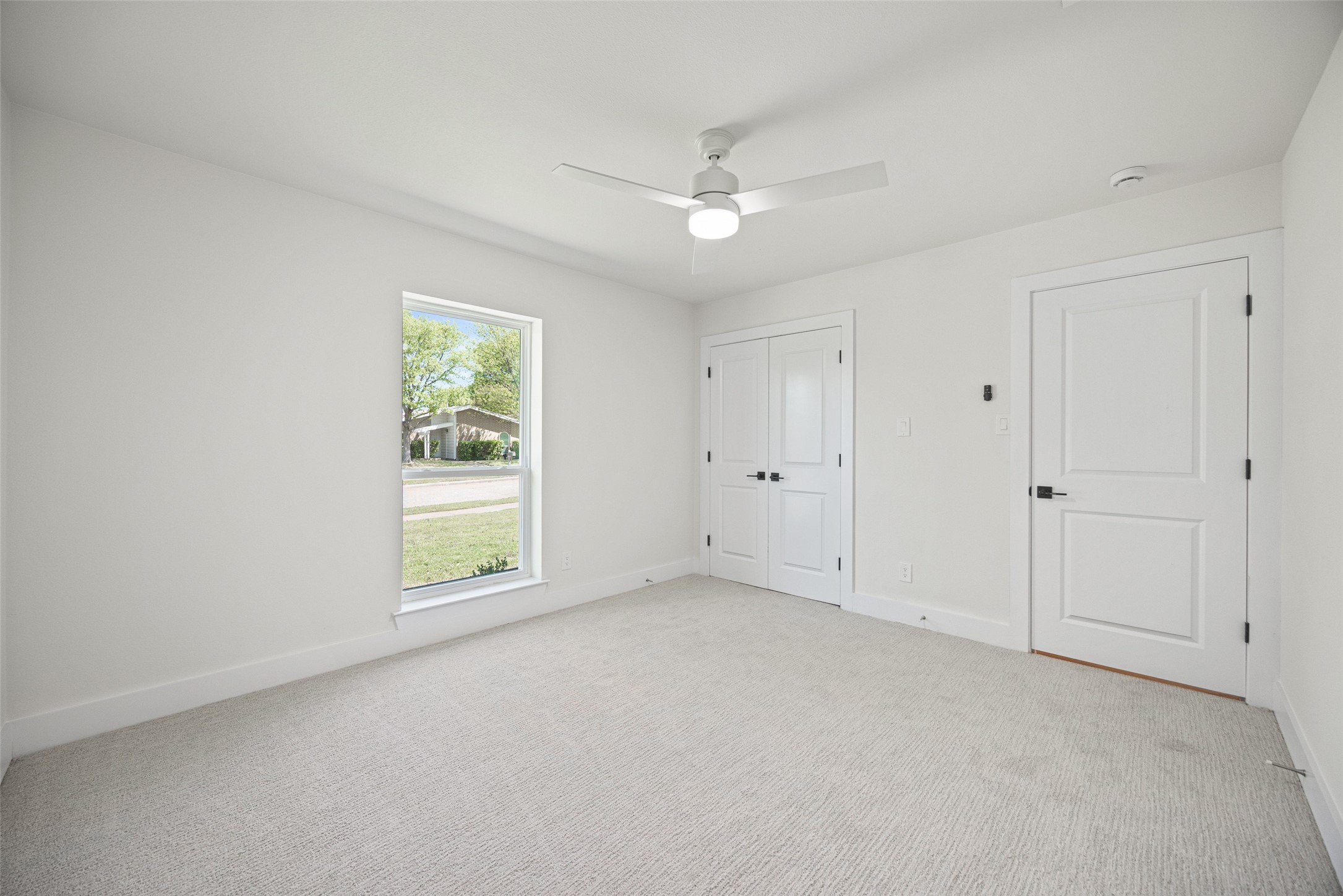 Vacant bedroom with light carpet, ceiling fan, large window, and double closet doors.