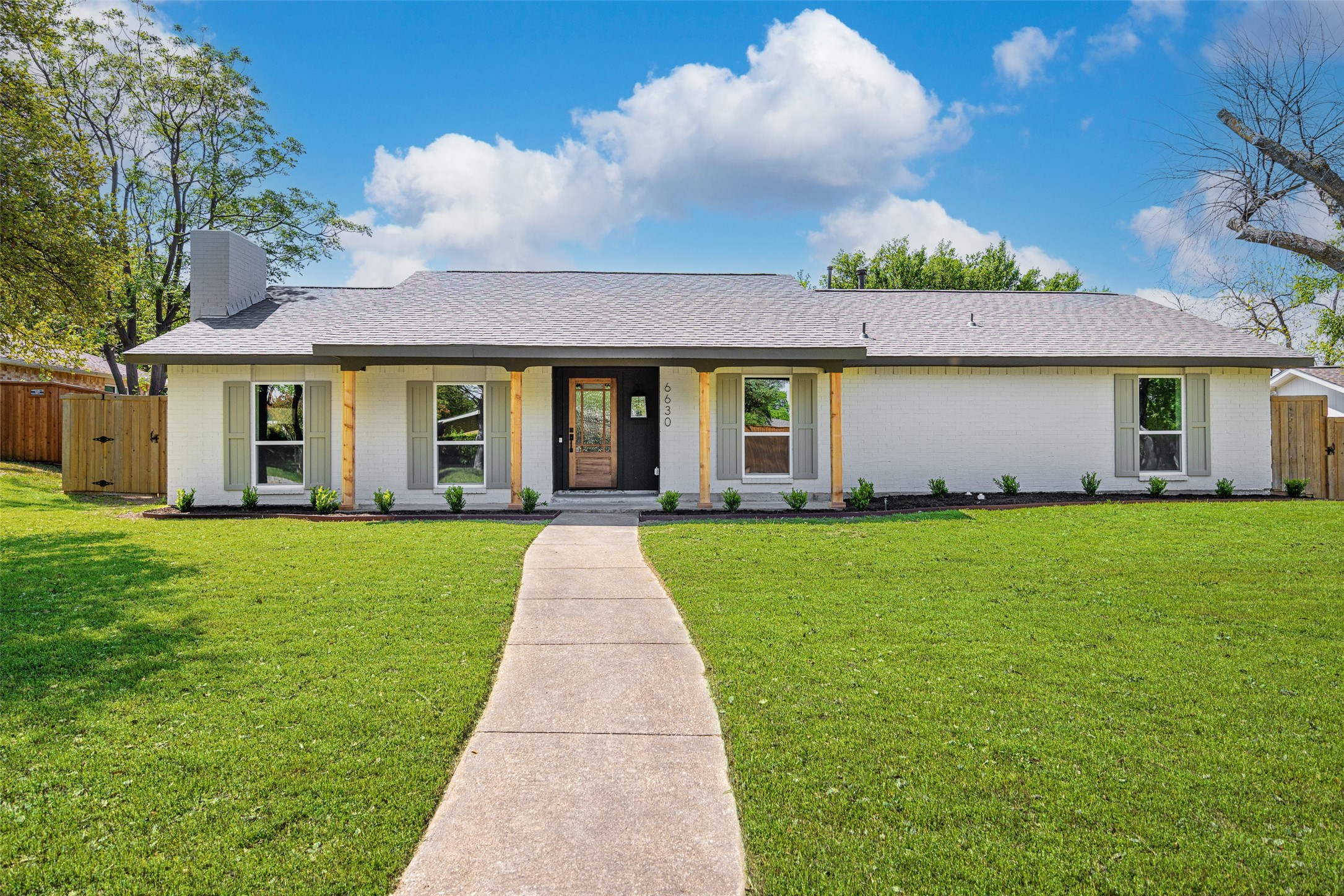 Single-story white brick home with a black entryway, shuttered windows, and curved walkway across a green lawn.