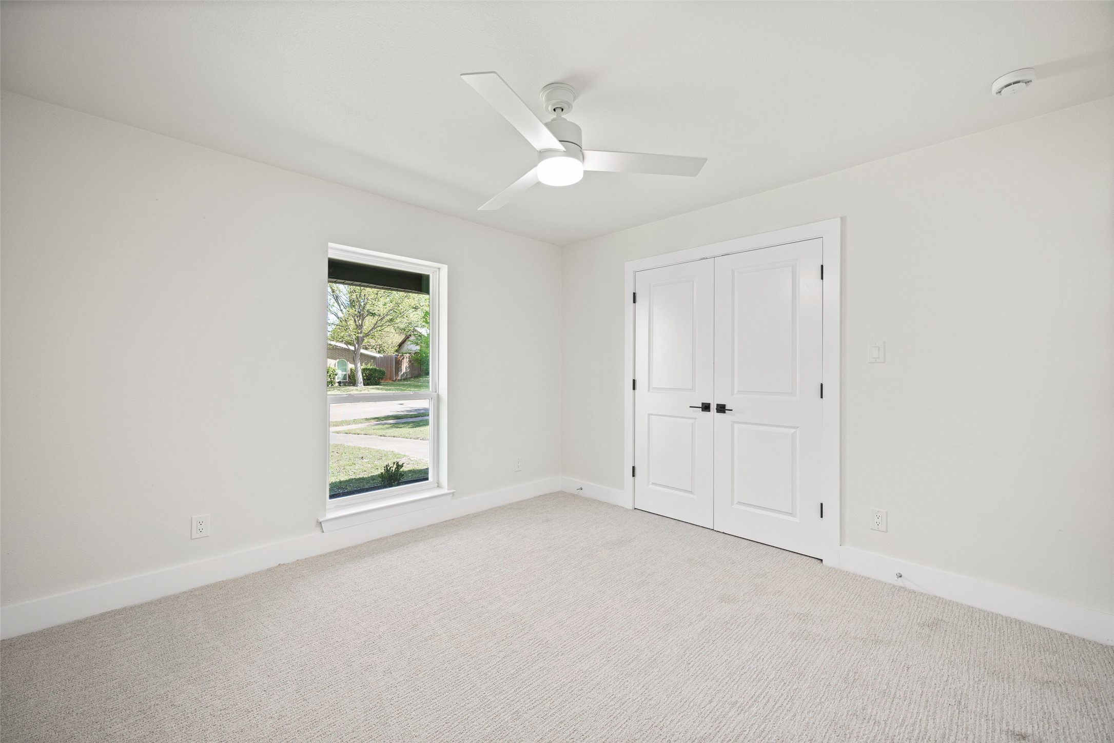 Empty carpeted bedroom with a ceiling fan, double closet doors, and a street-facing window.