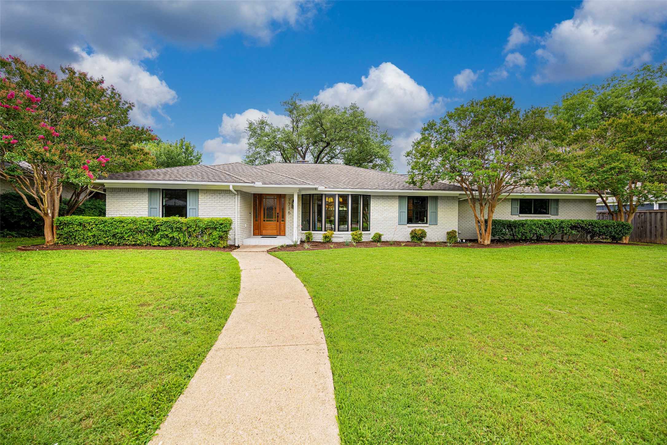 Single-story white brick home with a curved front walkway, broad lawn, and flowering trees.