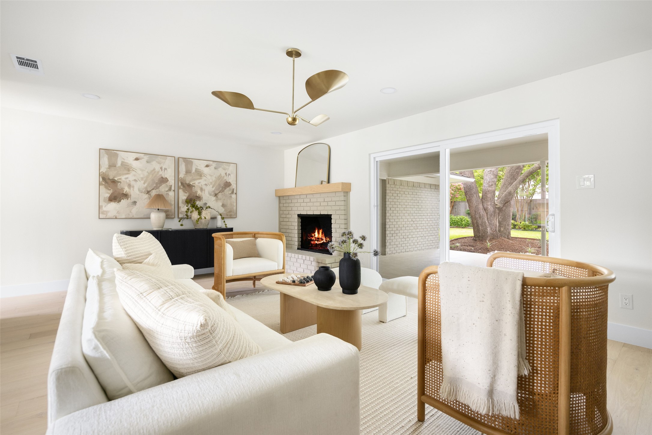Living room with a lit brick fireplace, white sofas, cane chairs, and doors opening to a leafy backyard.