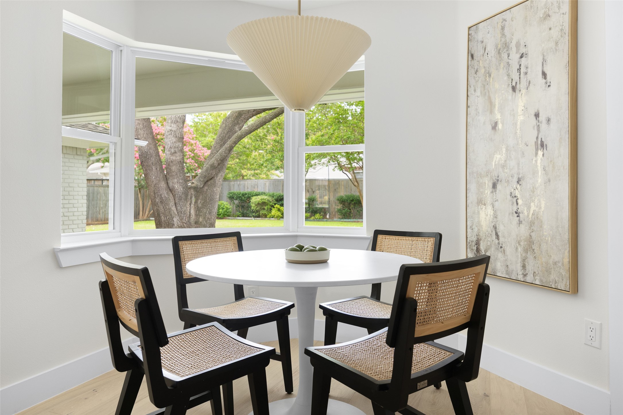 Dining nook with a round white table, woven chairs, and large windows overlooking a leafy backyard.