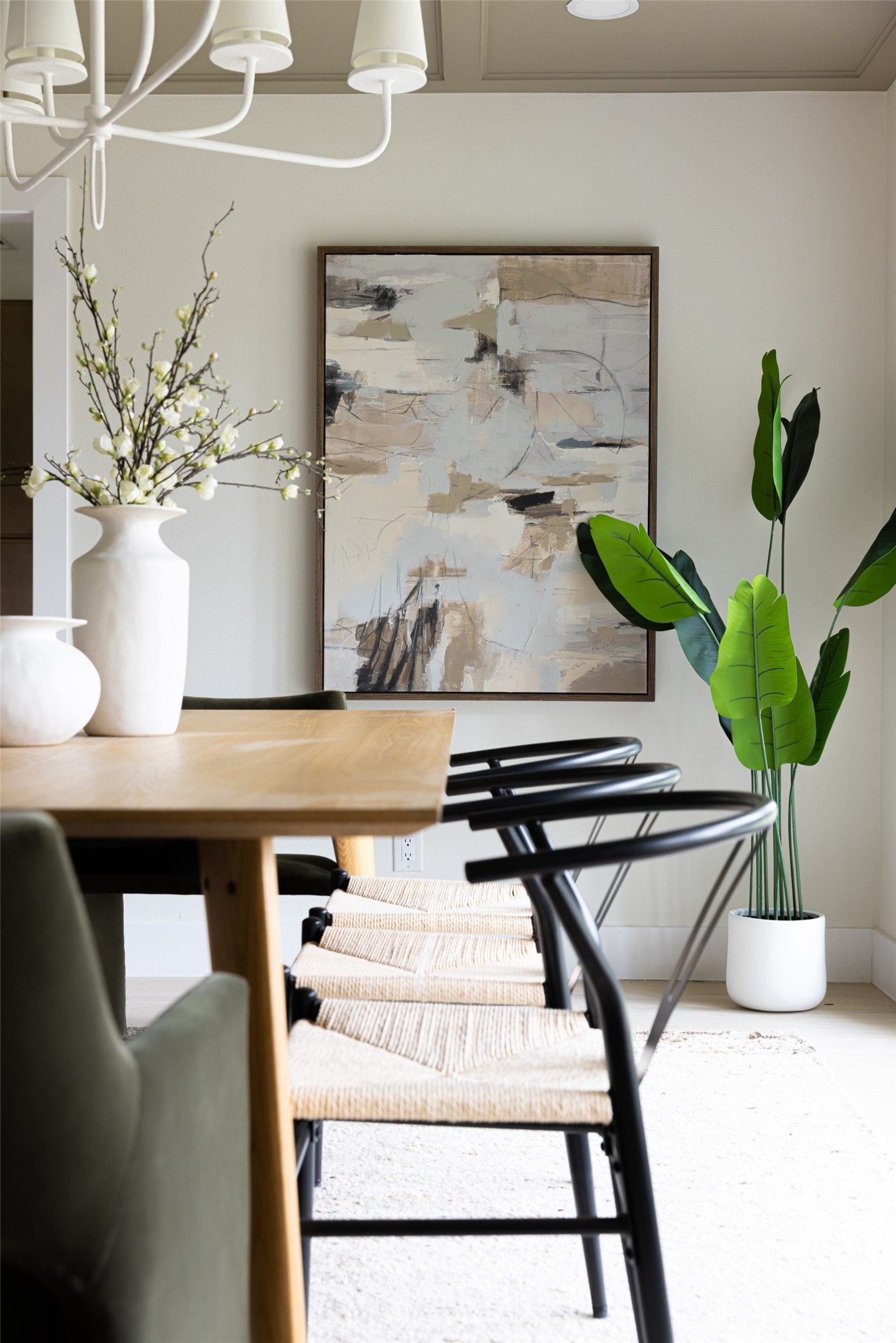 Dining room with a light wood table, woven chairs, abstract wall art, and a tall potted plant.