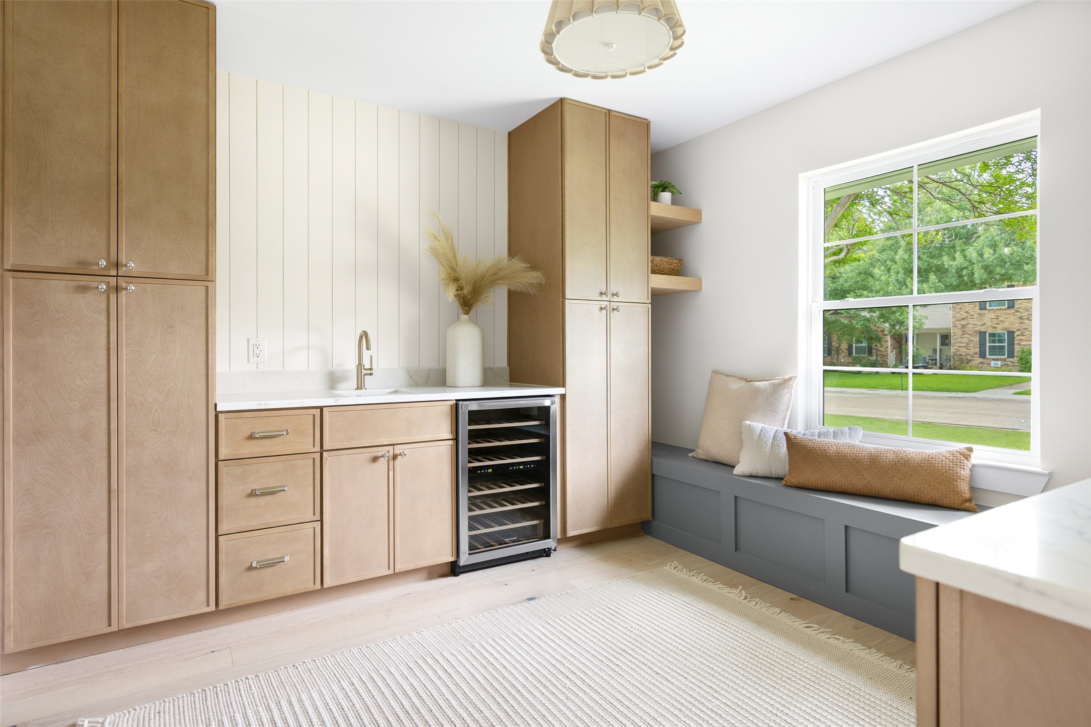 Kitchen beverage nook with light wood cabinets, wine refrigerator, and cushioned window seat beside a large window.