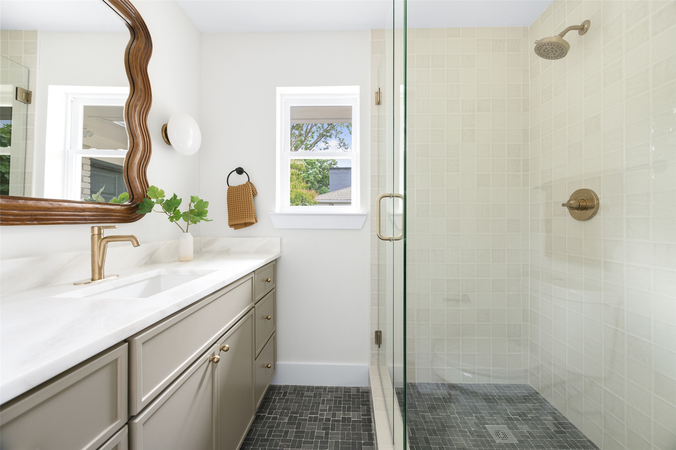 Bathroom with a marble-look vanity, wavy wood-framed mirror, and glass shower with brass fixtures.