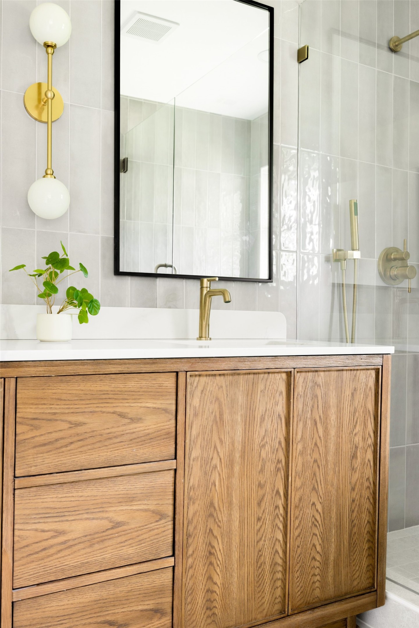 Bathroom vanity with a wood cabinet, brass fixtures, black-framed mirror, and glass shower enclosure.