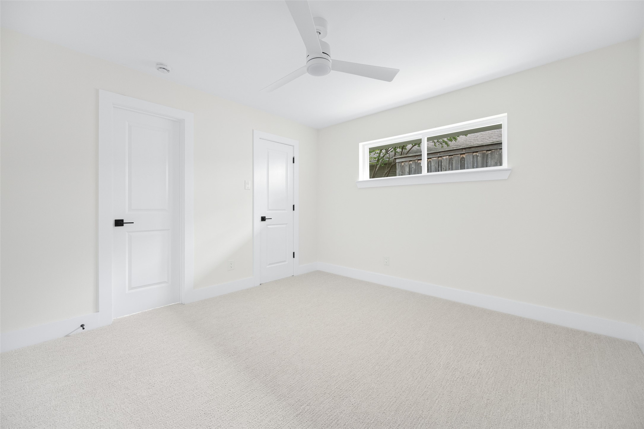 Empty bedroom with light carpet, a ceiling fan, and a narrow window overlooking a wooden fence.