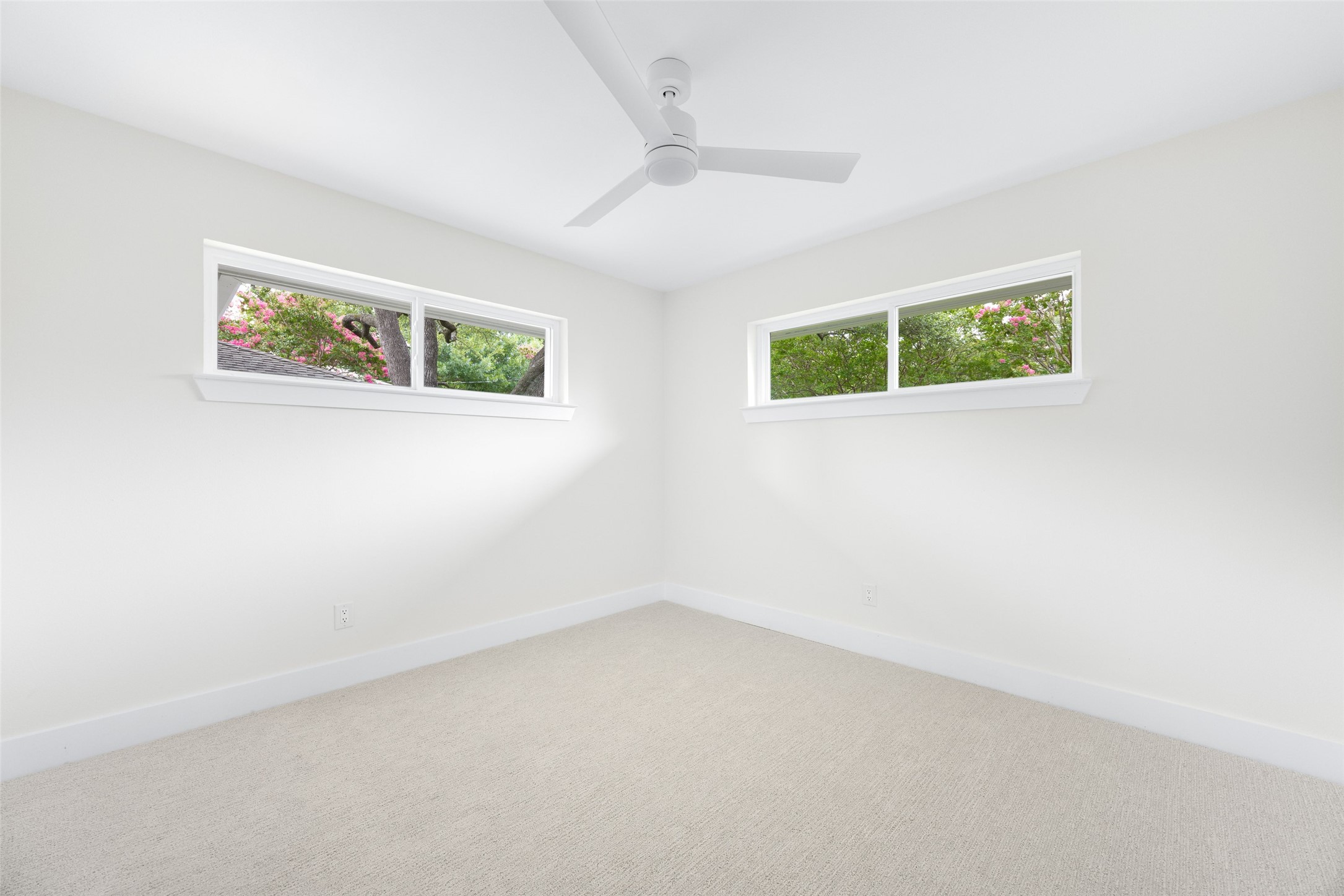 Vacant bedroom with a ceiling fan, beige carpet, and high windows overlooking flowering trees.