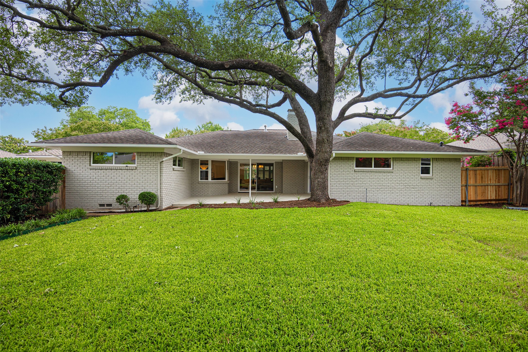 Single-story brick home with a covered patio beneath a large oak tree and a broad green lawn.