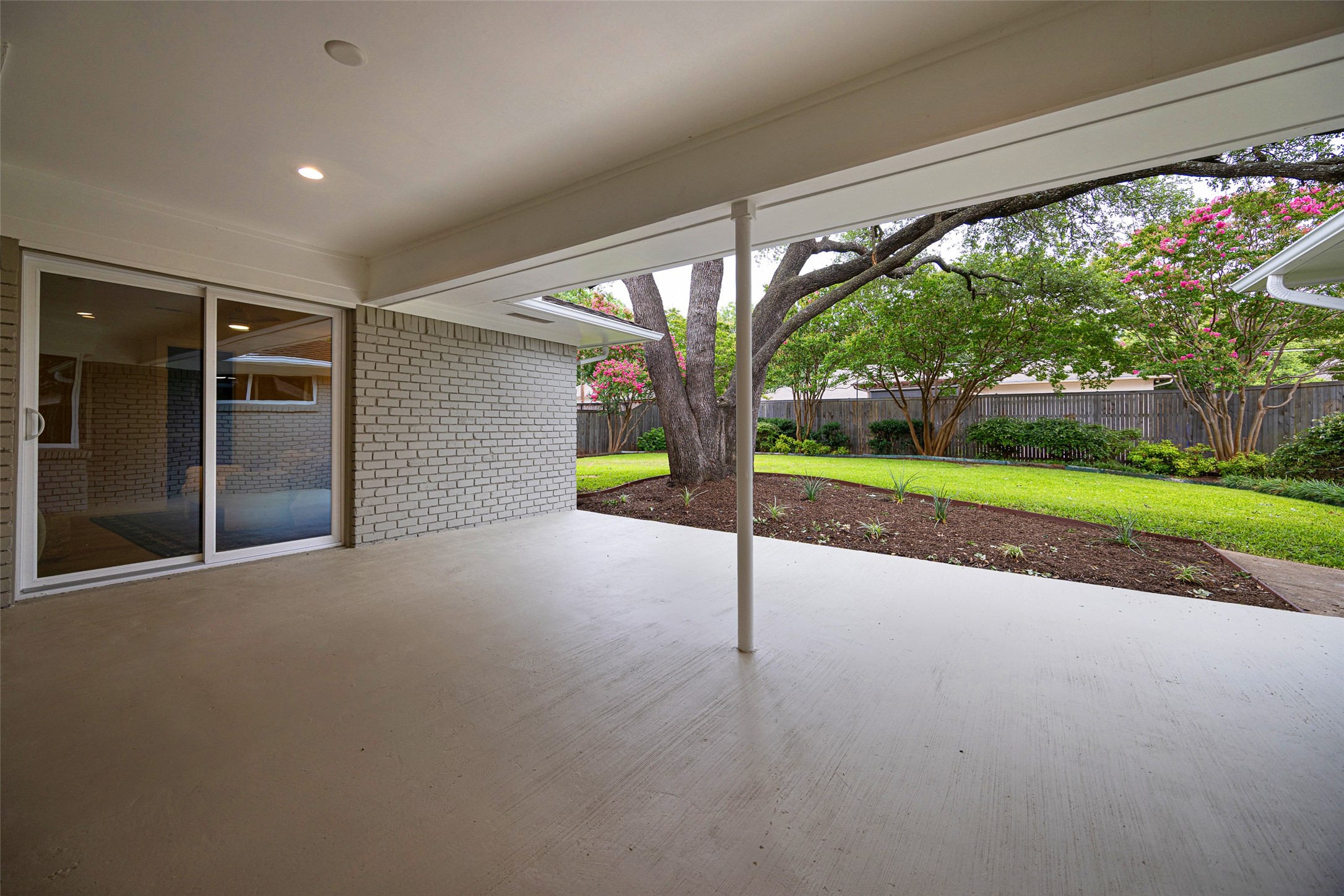 Covered concrete patio overlooking a landscaped backyard with mature trees and pink flowering crepe myrtles.