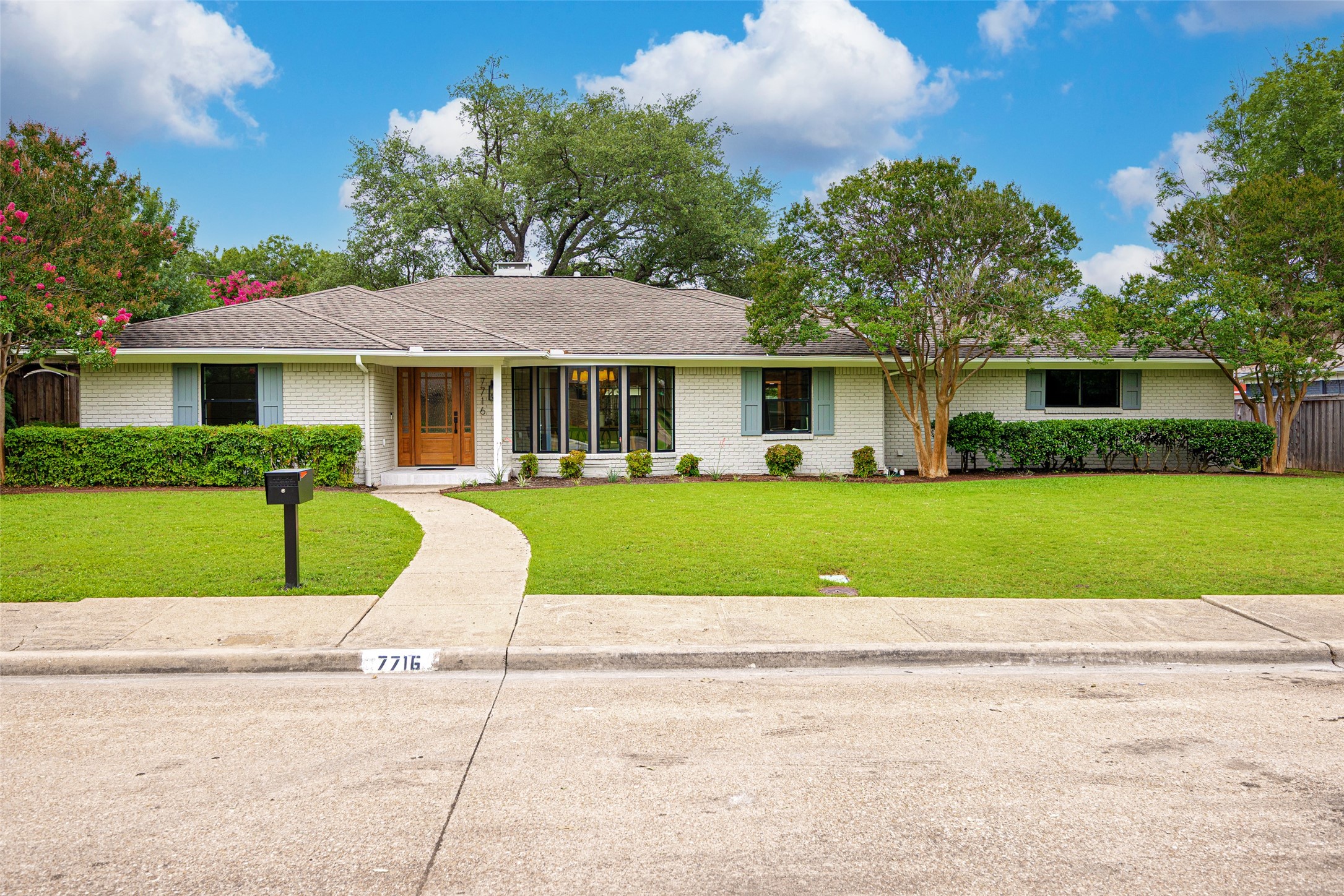 Single-story white brick home with a curved front walkway, green lawn, and mature trees.