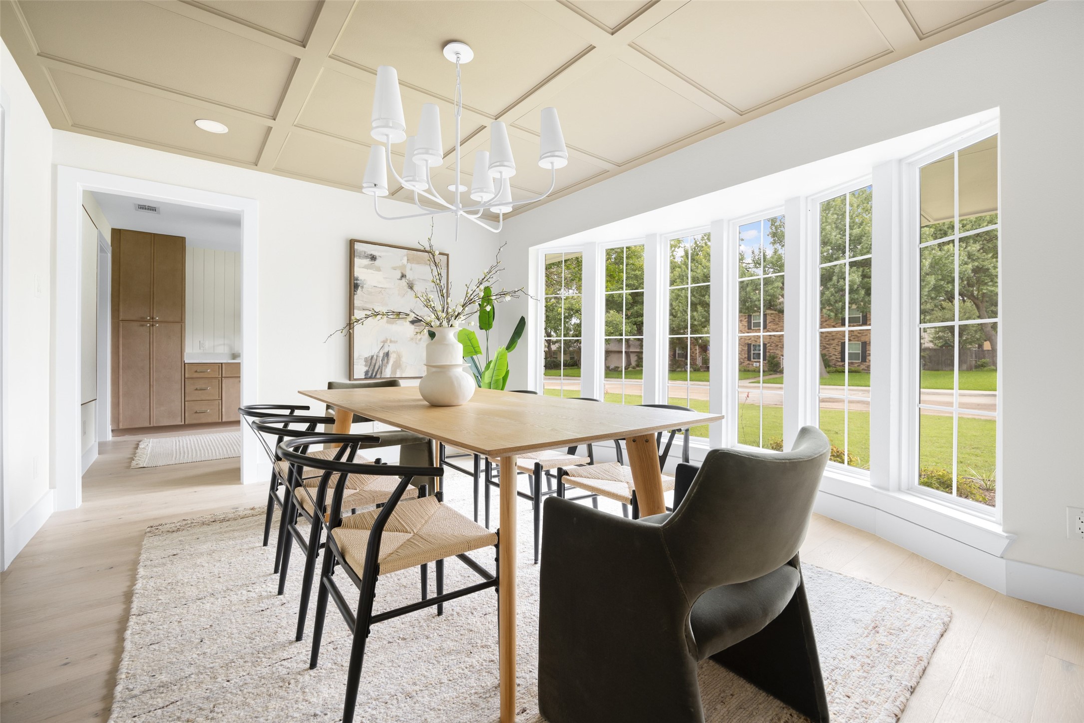 Dining room with a long wooden table, coffered ceiling, and tall windows overlooking a lawn.