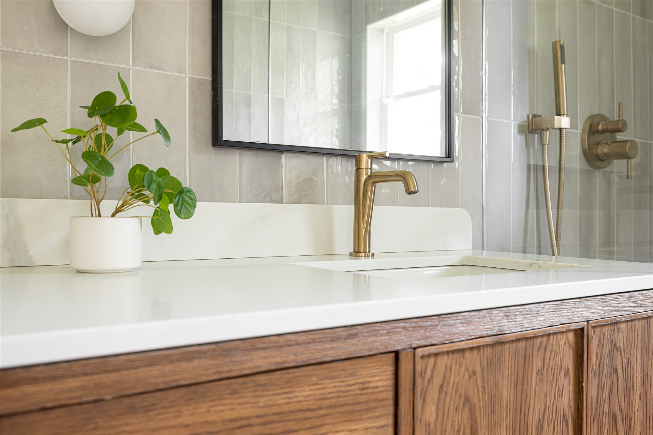 Bathroom vanity with a wood cabinet, brass fixtures, gray tile, and a potted plant.