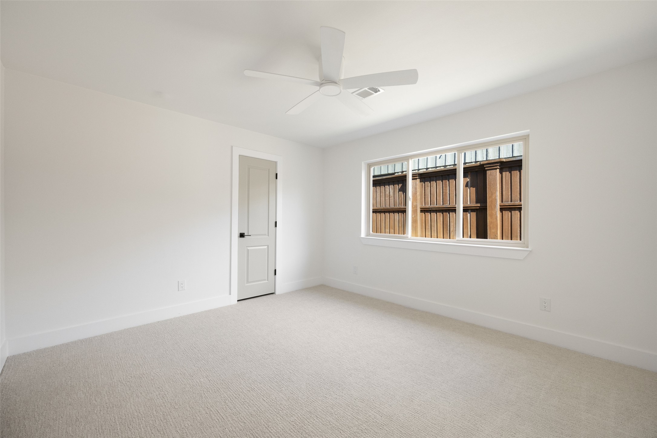 Empty bedroom with light carpet, a ceiling fan, and a wide window facing a wooden fence.
