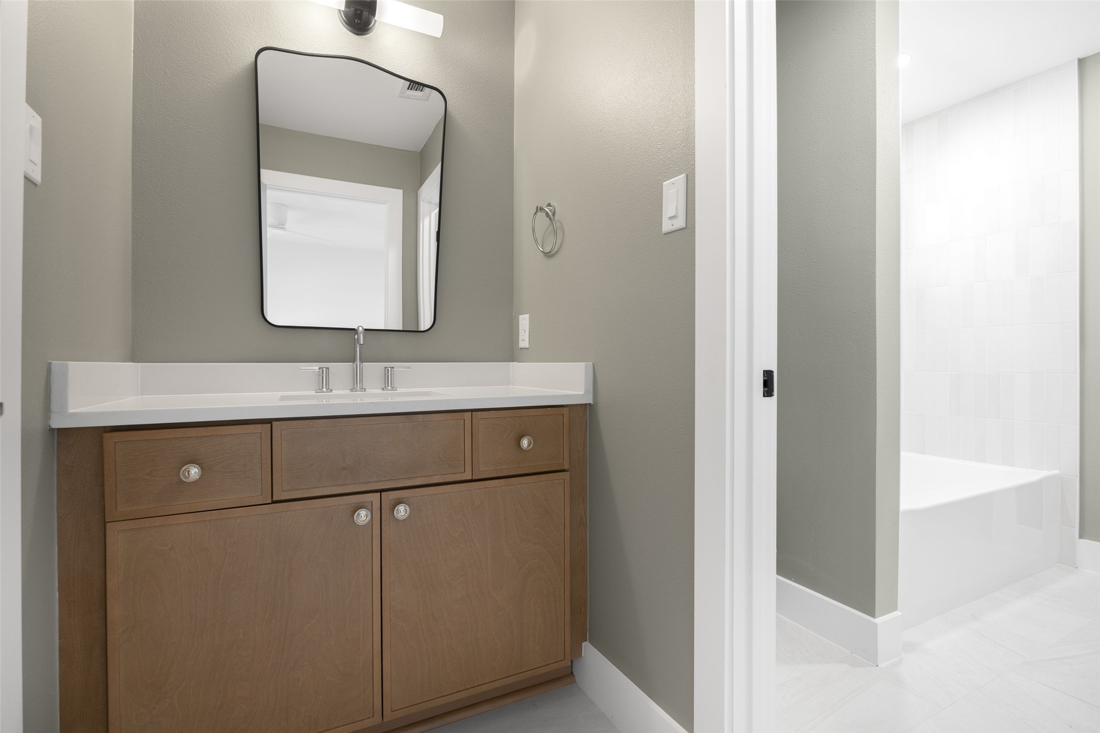 Bathroom vanity with a wood cabinet and mirror beside a white-tiled bathtub.