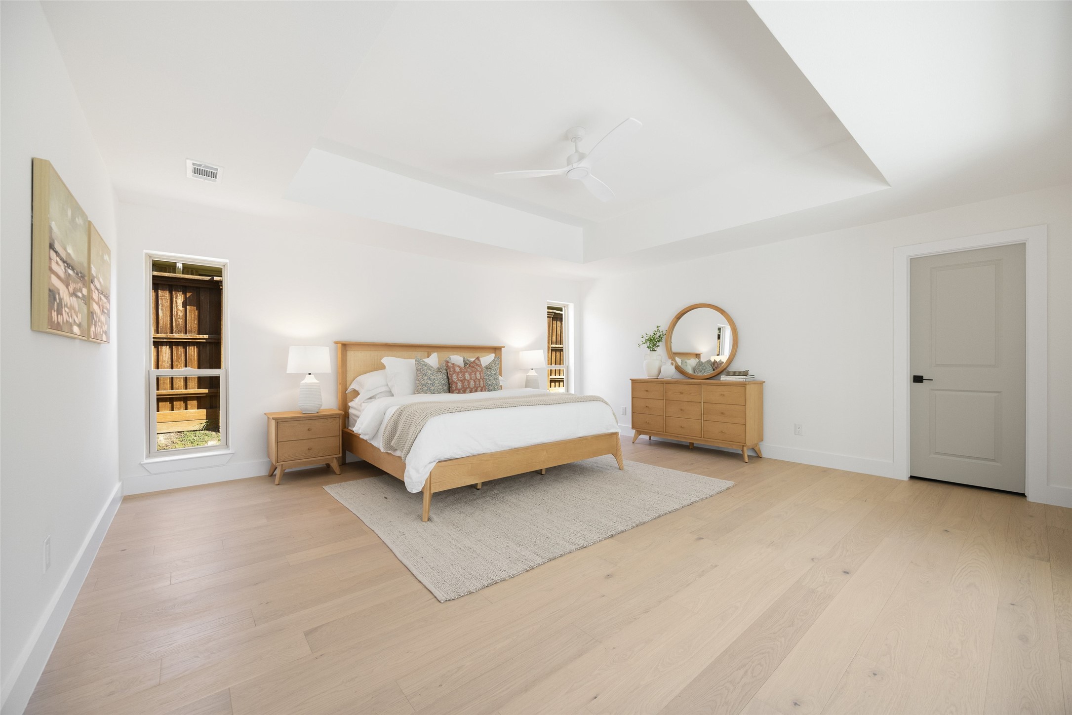 Staged bedroom with a king bed, light wood furniture, and a tray ceiling with a ceiling fan.
