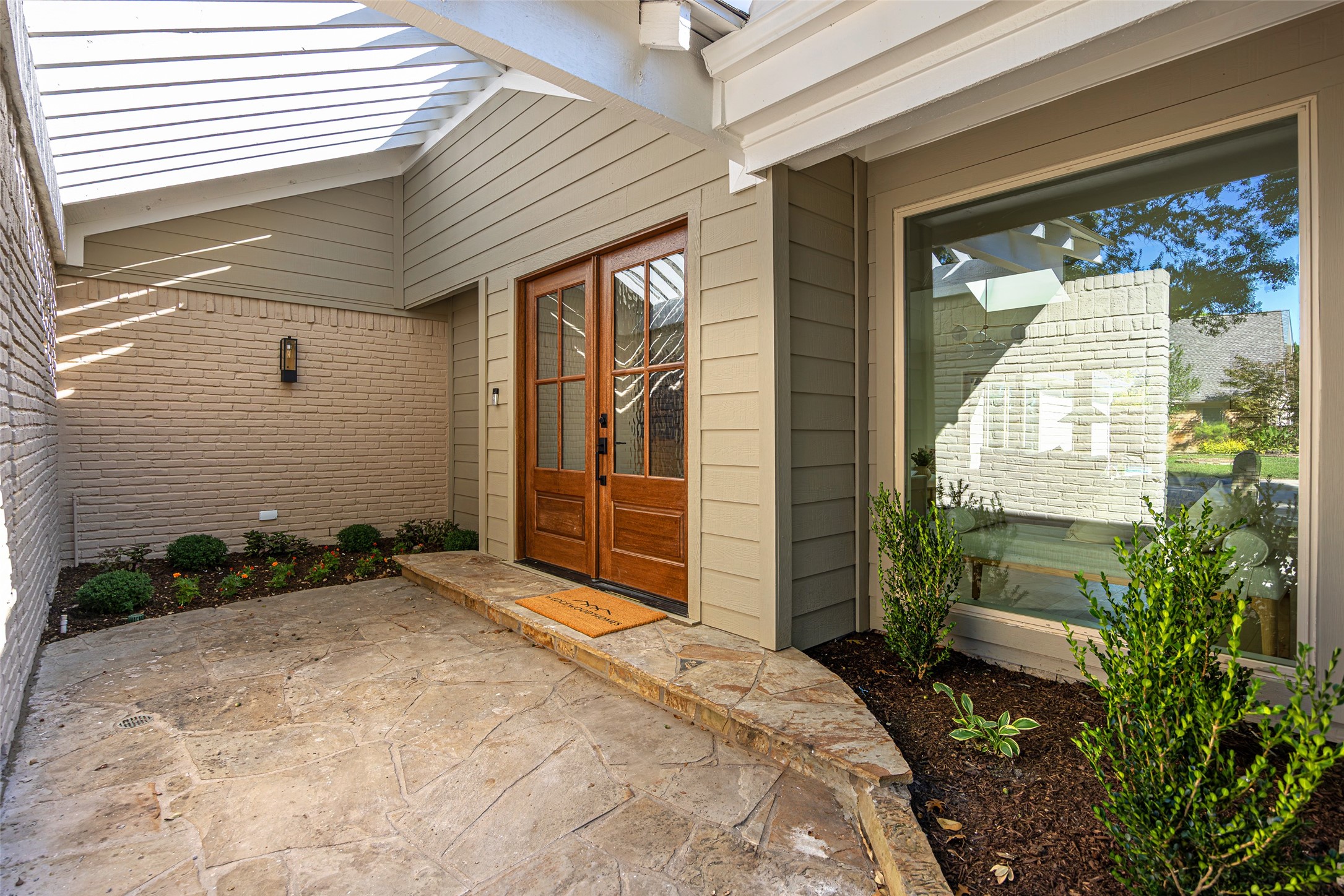 Covered stone-paved entry with wood double doors and landscaped planting beds.