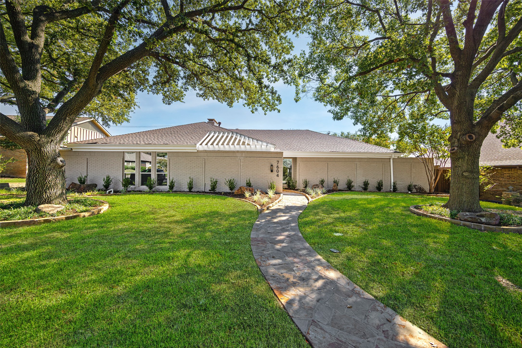 Single-story light brick home framed by mature trees, with a curved stone walkway through the front lawn.