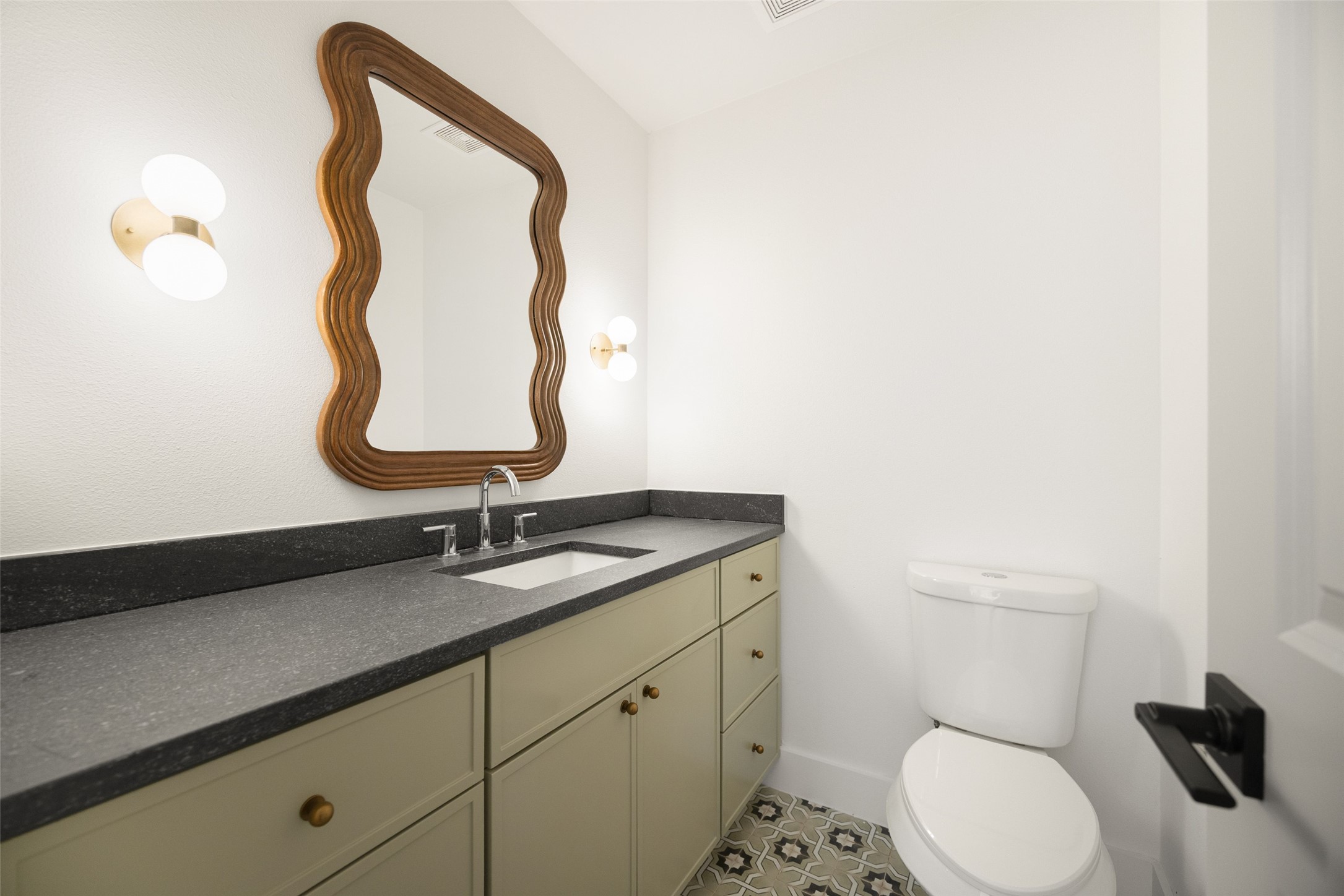 Bathroom with sage-green vanity, dark countertop, wavy wood-framed mirror, and patterned tile floor.
