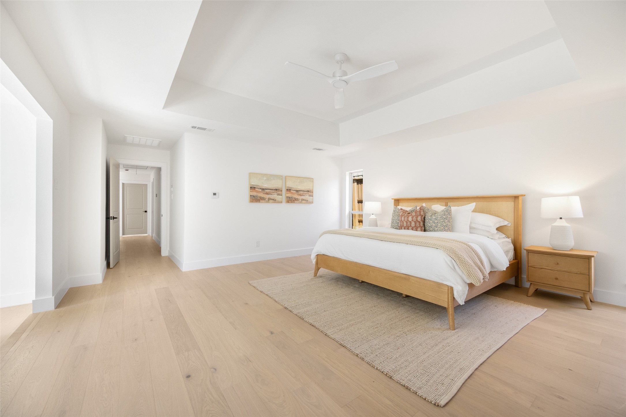 Staged bedroom with a wooden bed, white bedding, pale wood floors, and a tray ceiling with a fan.