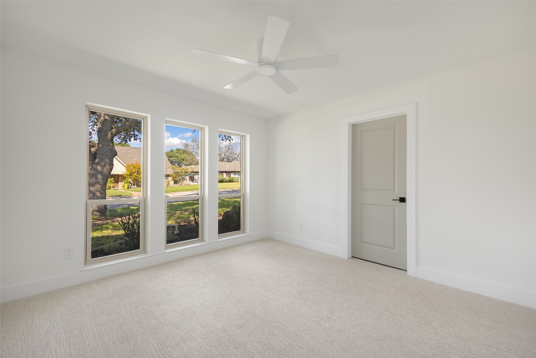 Empty carpeted room with a ceiling fan and three tall windows overlooking a tree-lined street.