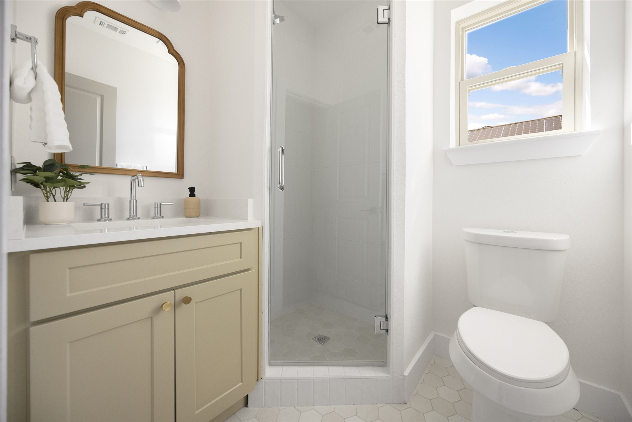 Bathroom with beige vanity, glass shower, hexagonal tile, and a toilet beside a bright window.