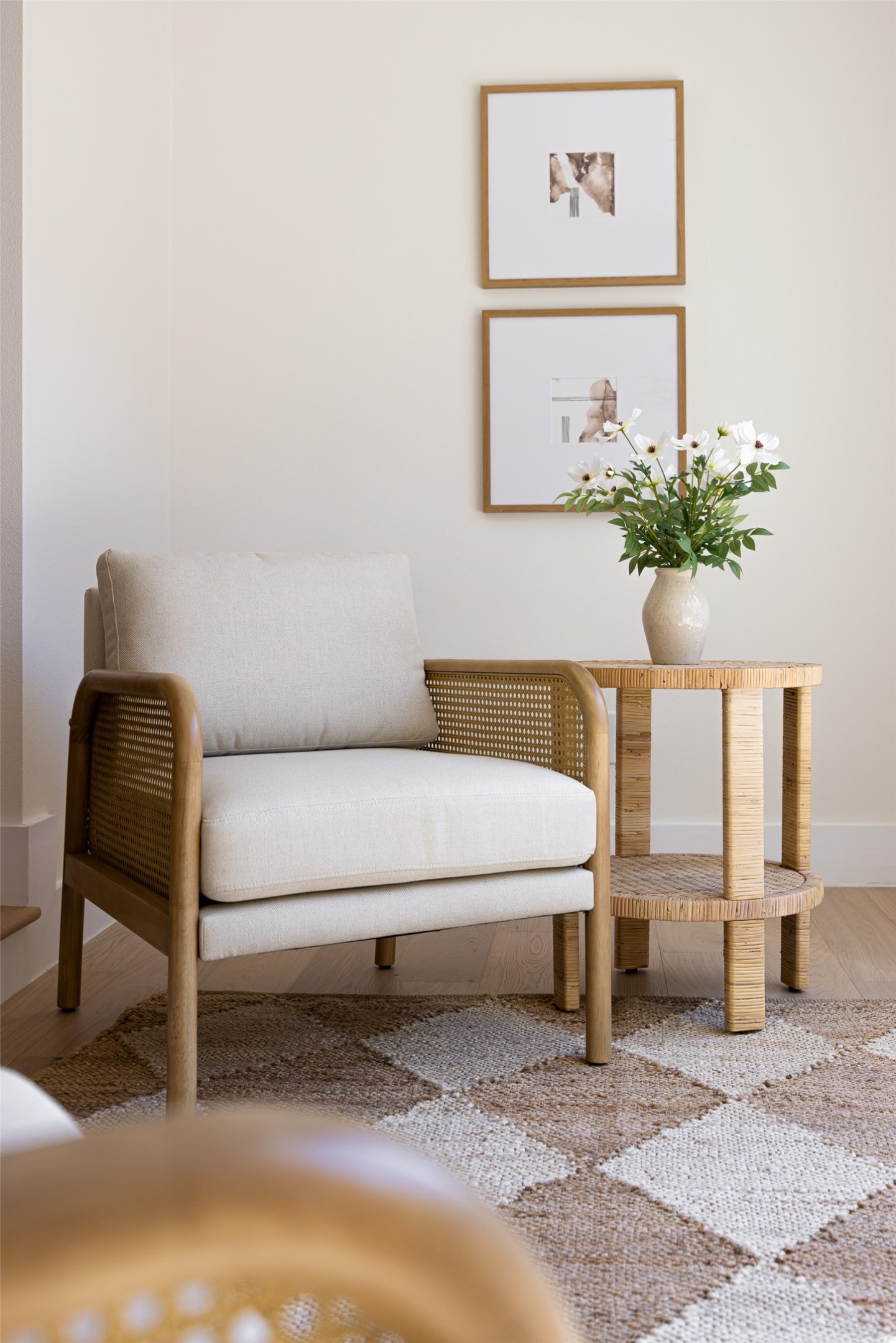 Cream chair with woven arms beside a round table of white flowers, against a pale wall with framed art.