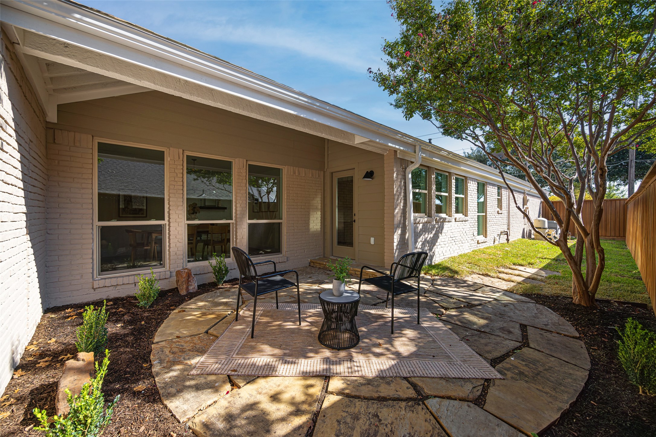 Rear patio with stone paving, two black chairs, and a mature tree beside a brick home.
