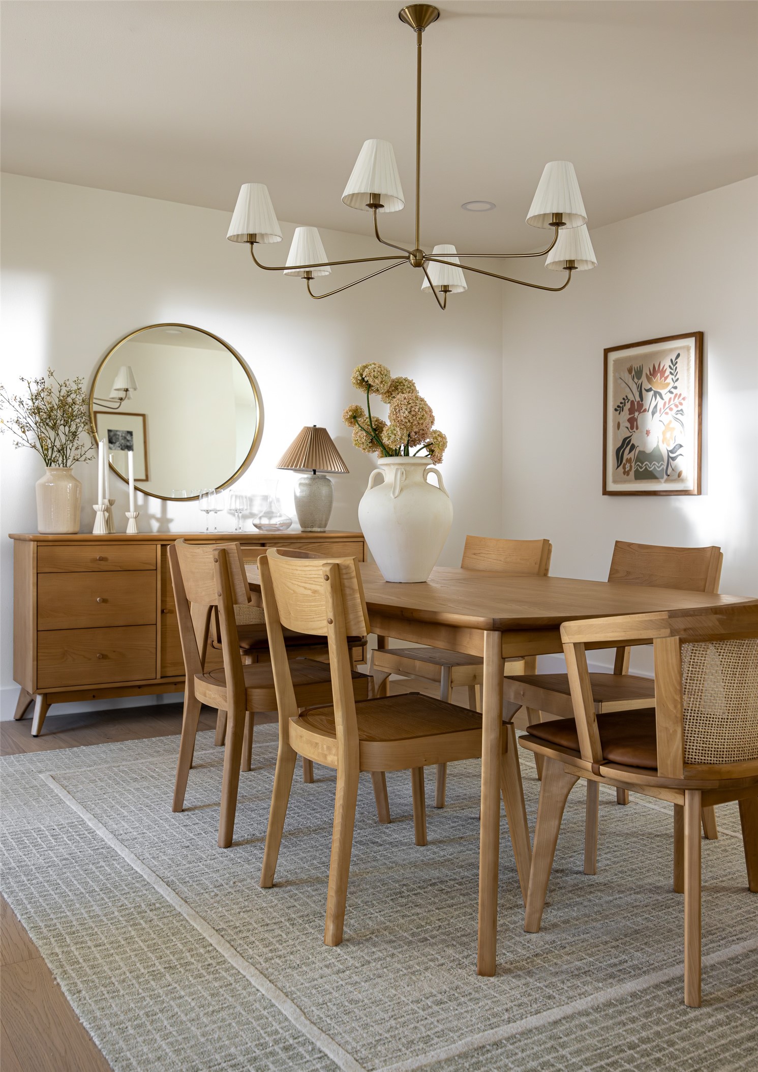 Dining room with a light wood table, brass chandelier, round mirror, and wooden sideboard.