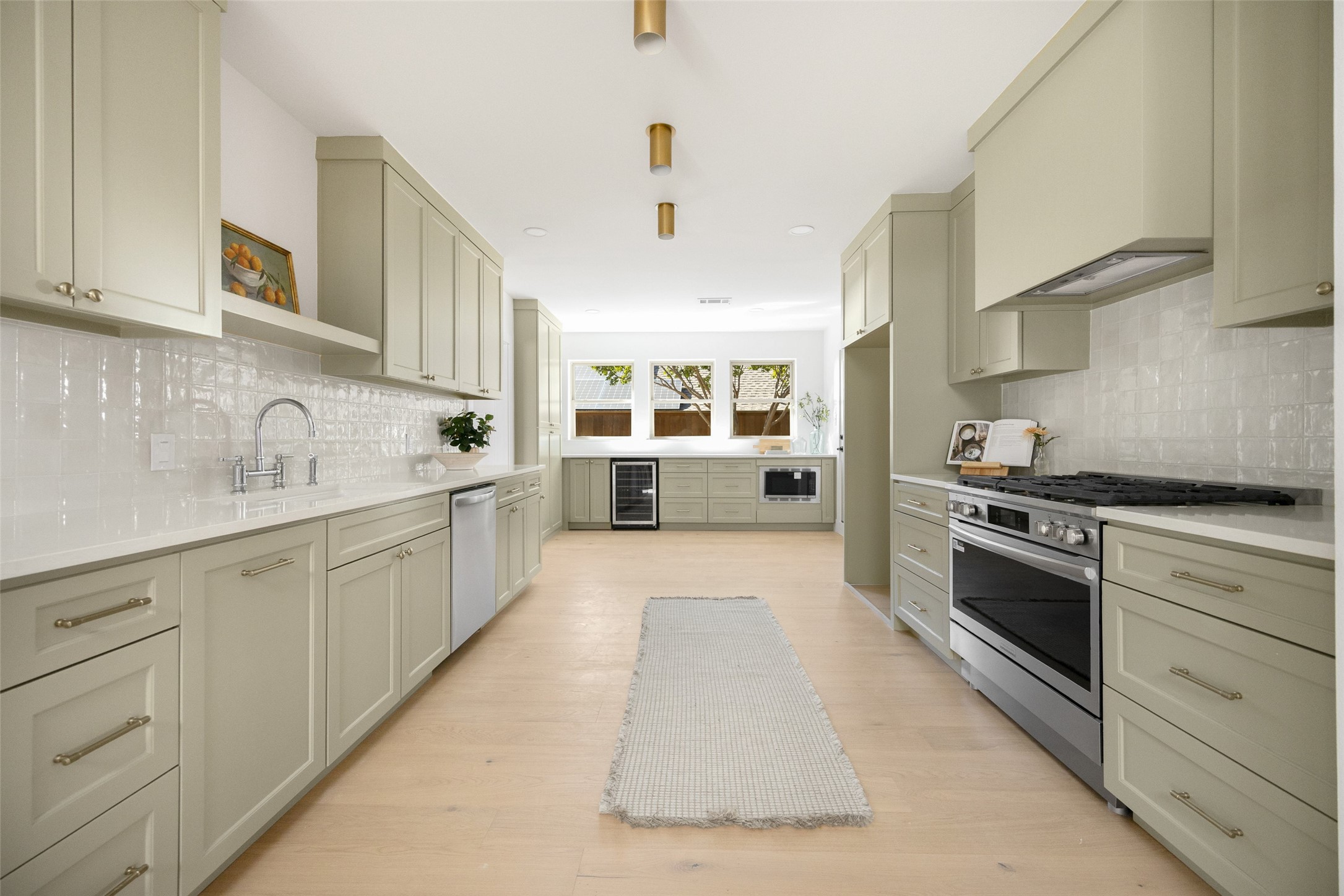 Bright galley kitchen with sage-gray cabinets, a stainless-steel range, and three windows at the far end.