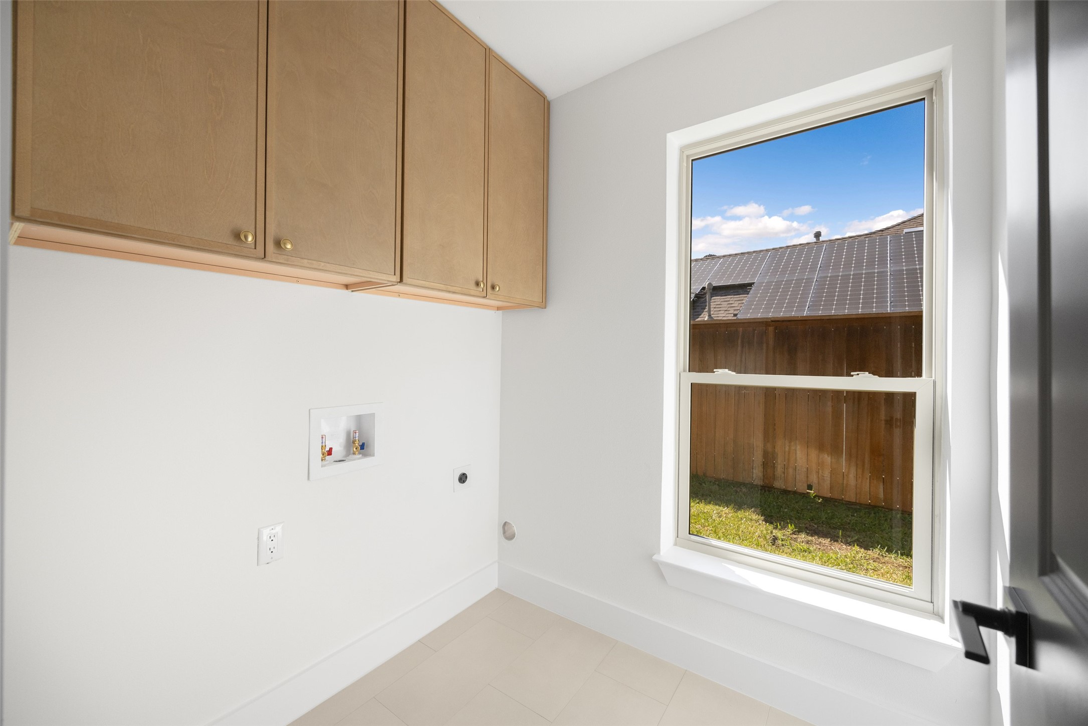 Empty laundry room with upper cabinets, appliance hookups, and a window overlooking a fenced yard.