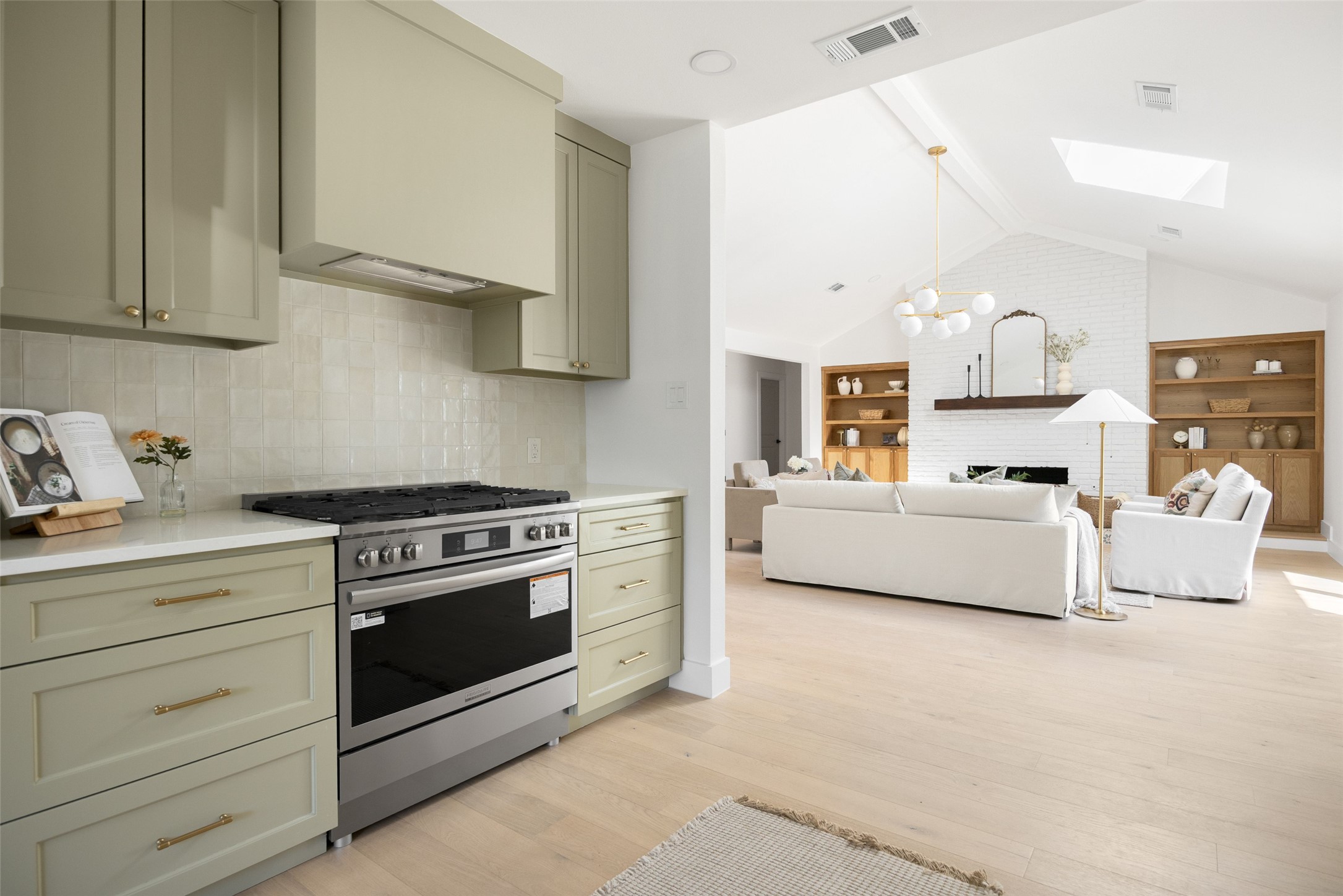 Open kitchen and living room with sage cabinets, a stainless range, white sofas, and a brick fireplace.