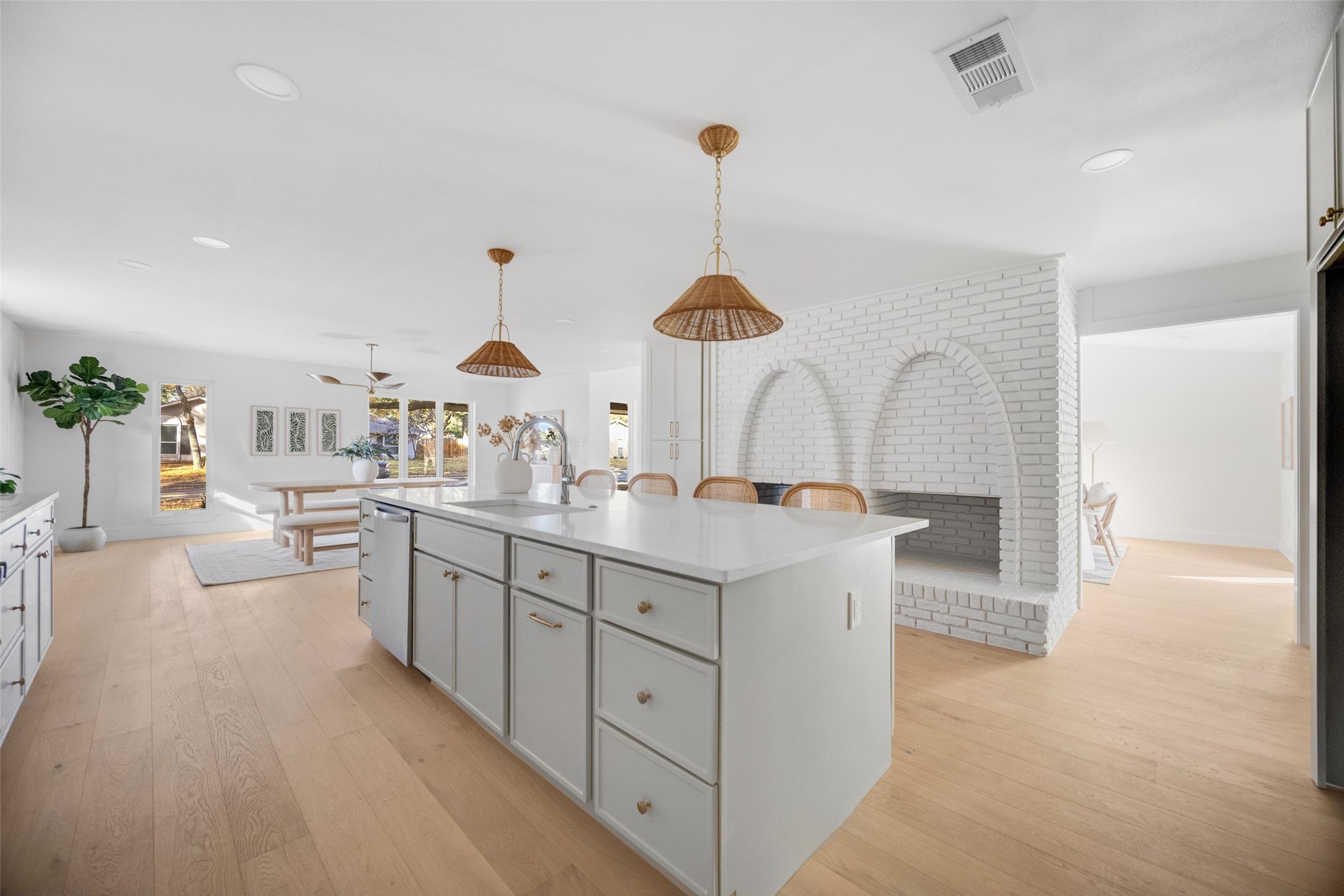 Open-plan kitchen with a large white island, woven pendant lights, and a white brick fireplace.