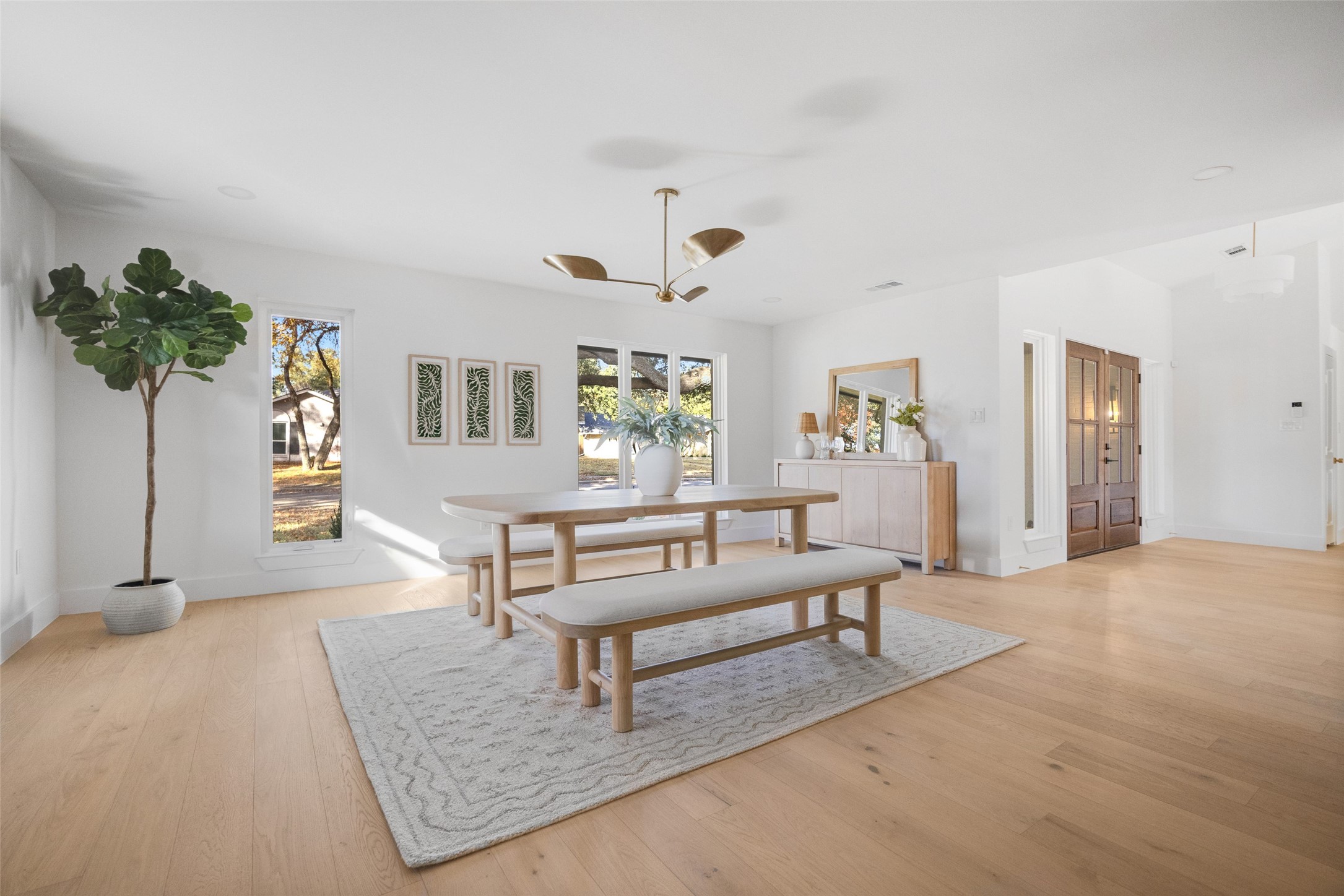 Dining room with a light wood table, upholstered benches, large windows, and double wooden entry doors.