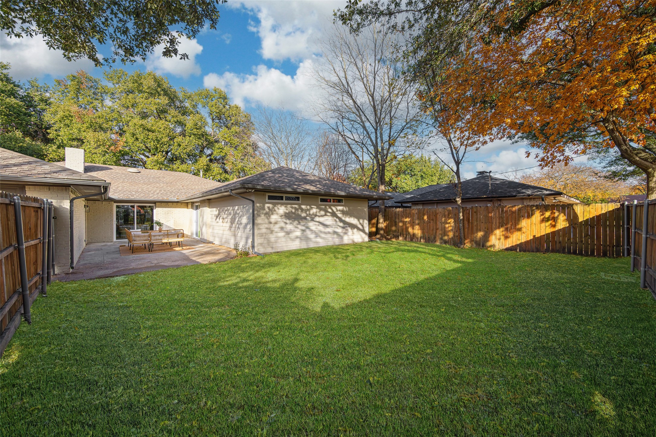 Fenced backyard with a broad lawn, covered patio seating, and mature autumn trees.